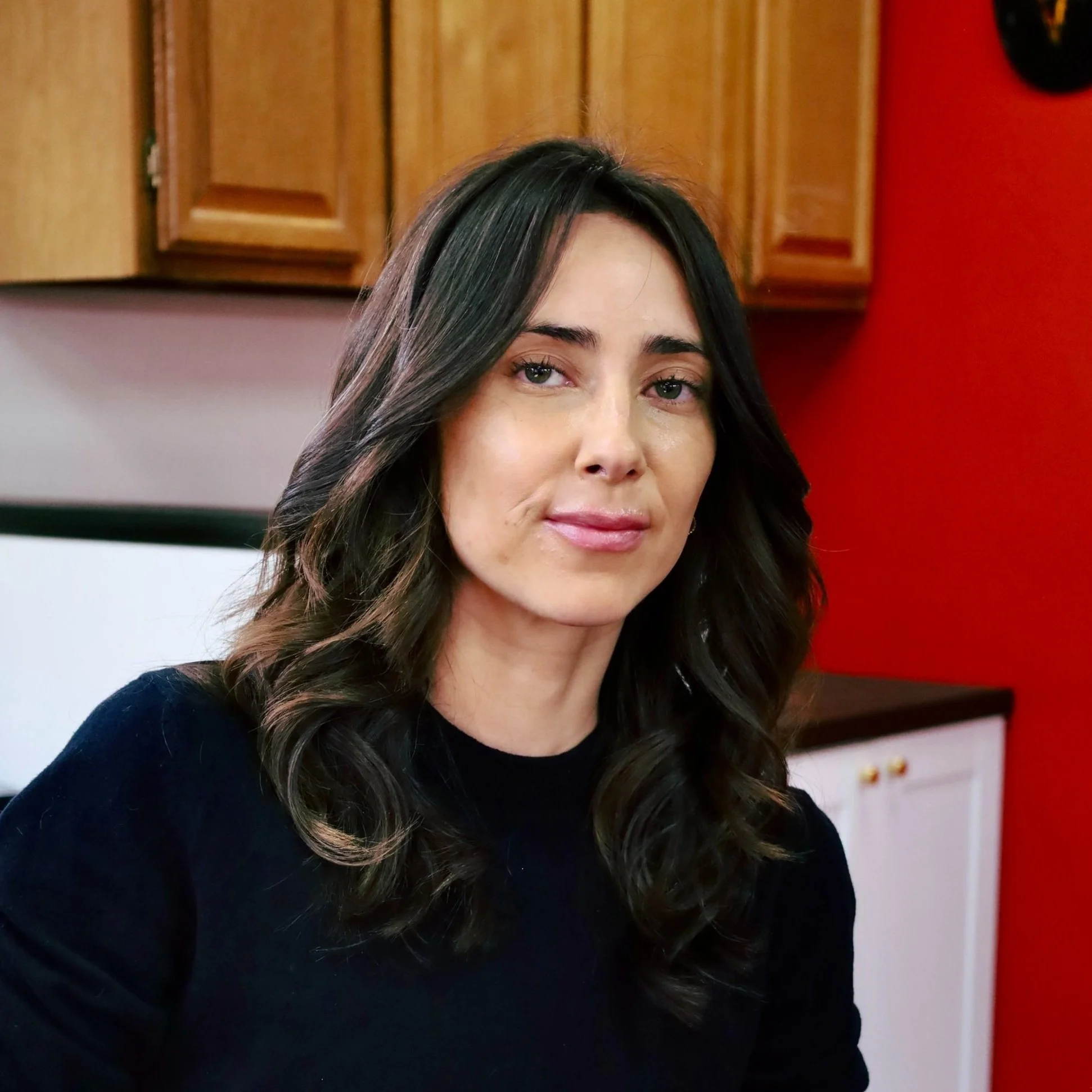 A woman with dark, wavy hair and light skin, wearing a black top, sitting indoors with wooden cabinets and a red wall in the background.