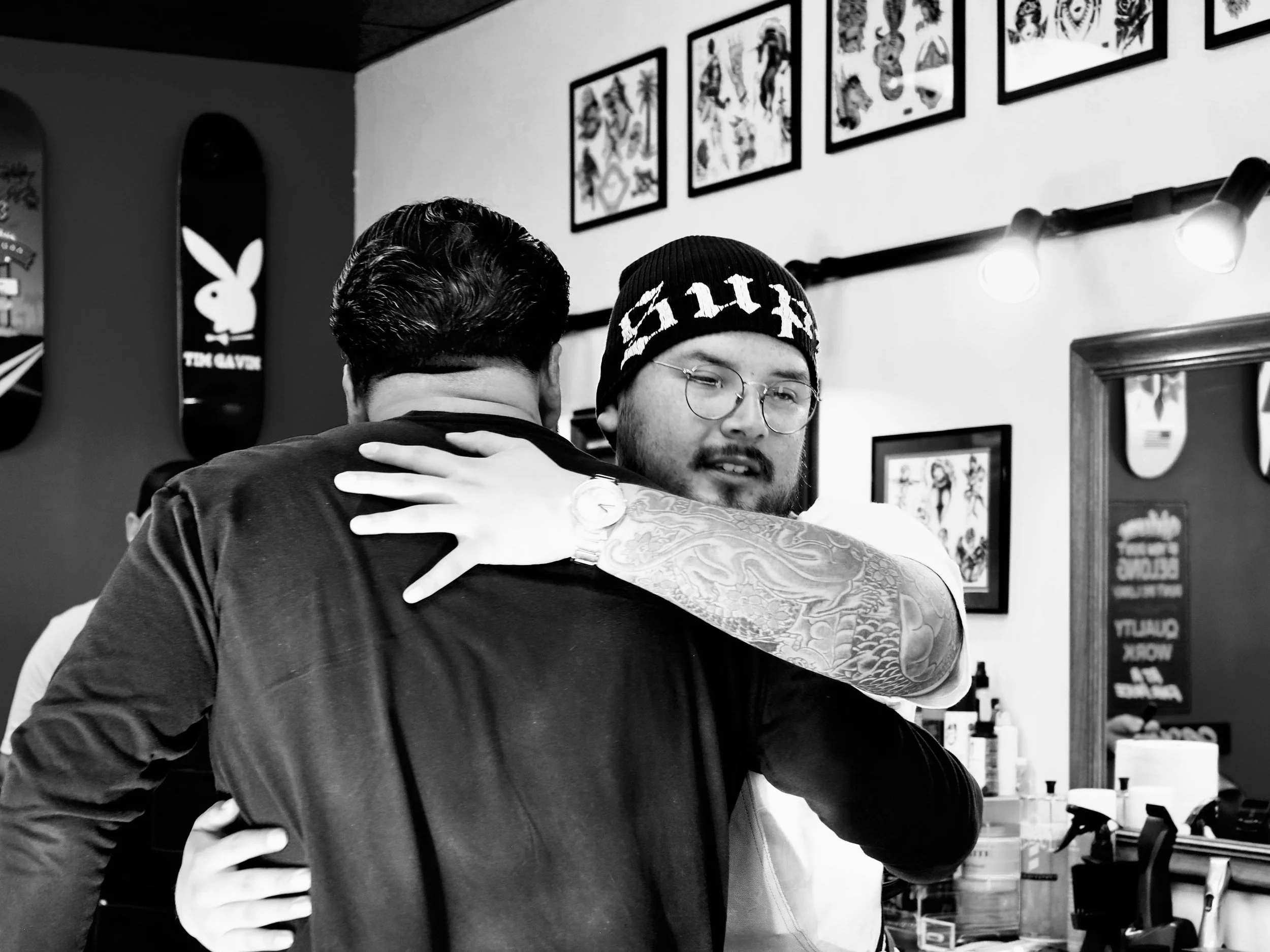 Two men hugging in a tattoo shop, one facing away and the other facing the camera, with framed artwork and skateboard decks on the walls.