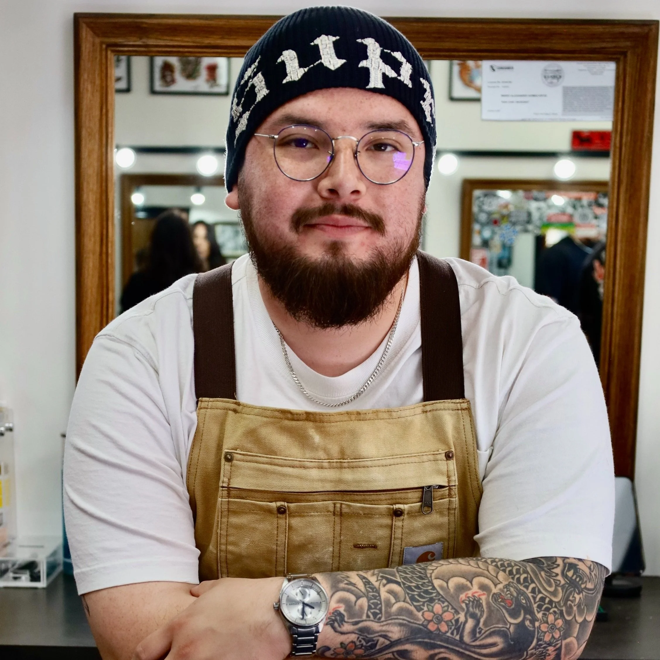 Barber Mario is a young man with glasses, a beard, and a tattooed arm, wearing a black beanie and a beige apron, standing inside a cafe or restaurant.
