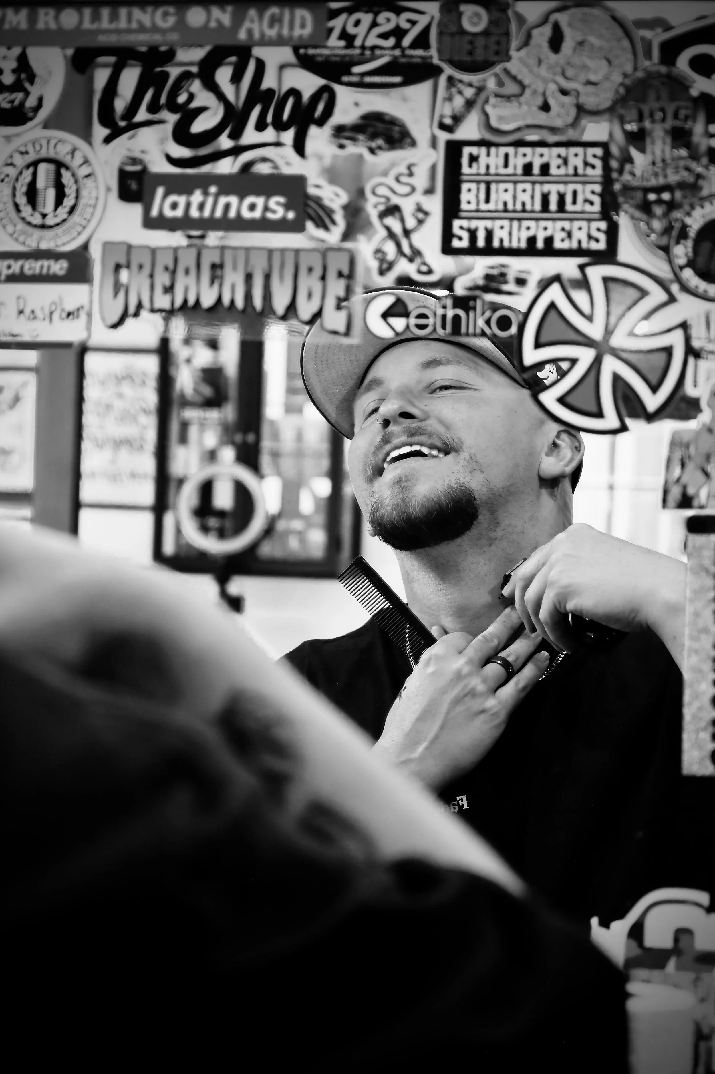 A man with a beard and mustache smiling and holding a pair of combs around his neck, surrounded by various stickers and posters on the wall behind him.