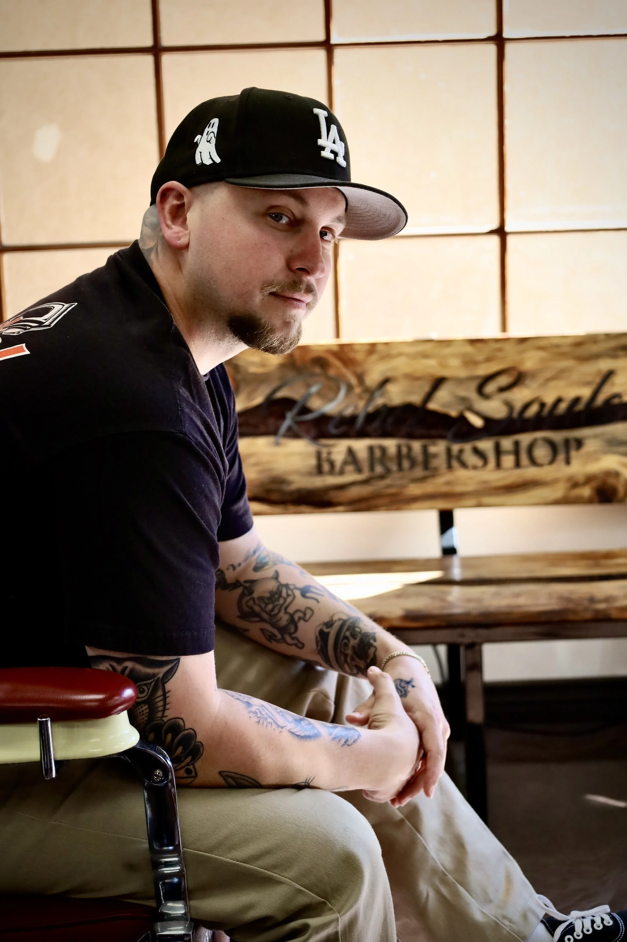 Austin Jacquemin, Owner + Barber at Rebel Souls Barbershop, a man with tattoos on his arms, wearing a black T-shirt and a baseball cap, sitting indoors near a wooden sign that reads 'Rebel Souls Barbershop'.