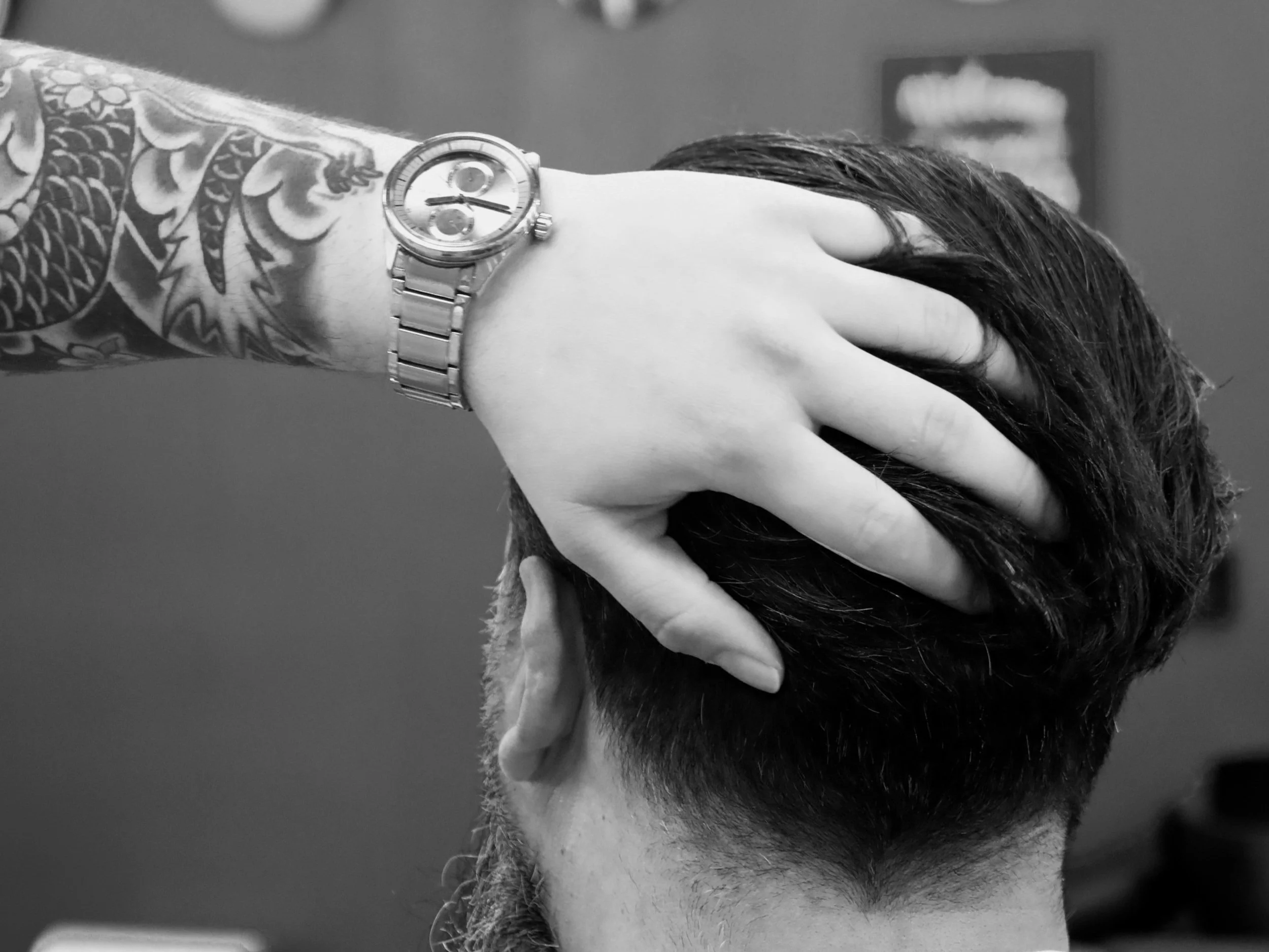 A person with a tattooed arm wearing a wristwatch, holding their head with their hand, showing short dark hair and part of their ear, in a black and white photo.