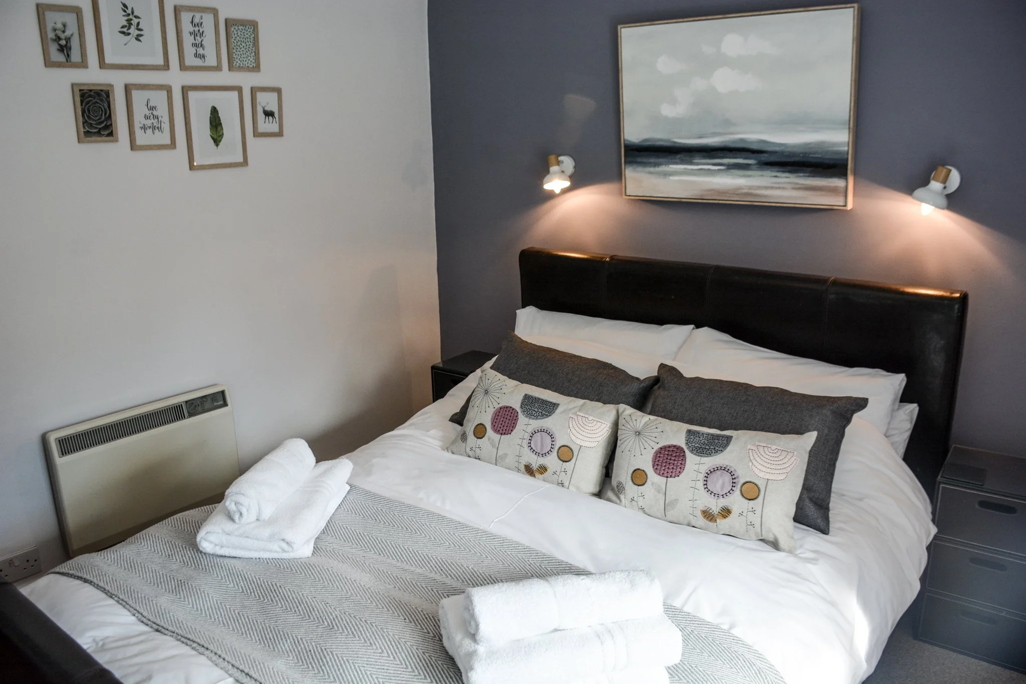 A neatly made bed with white bedsheets and decorated pillows, including two dark gray and three embroidered ones with colorful patterns. Rolls of white towels are placed at the foot of the bed. A large abstract landscape painting hangs above the bed, illuminated by two wall-mounted lights. To the left, there is a gallery wall with ten framed pictures and a small black side table on the right.