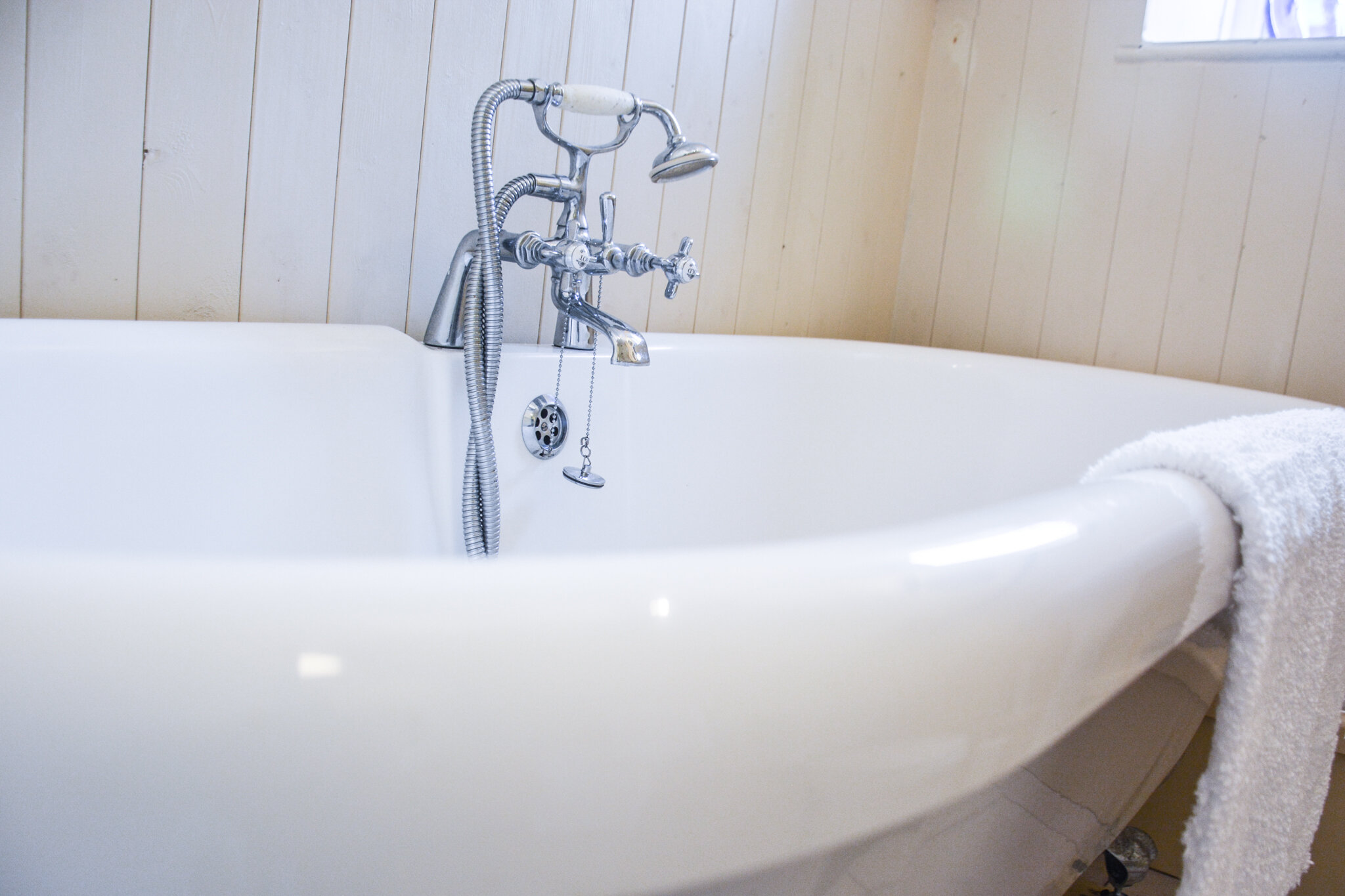 White vintage clawfoot bathtub with chrome fixtures, a handheld showerhead, and a white towel draped over the side, in a bathroom with wood-paneled walls.