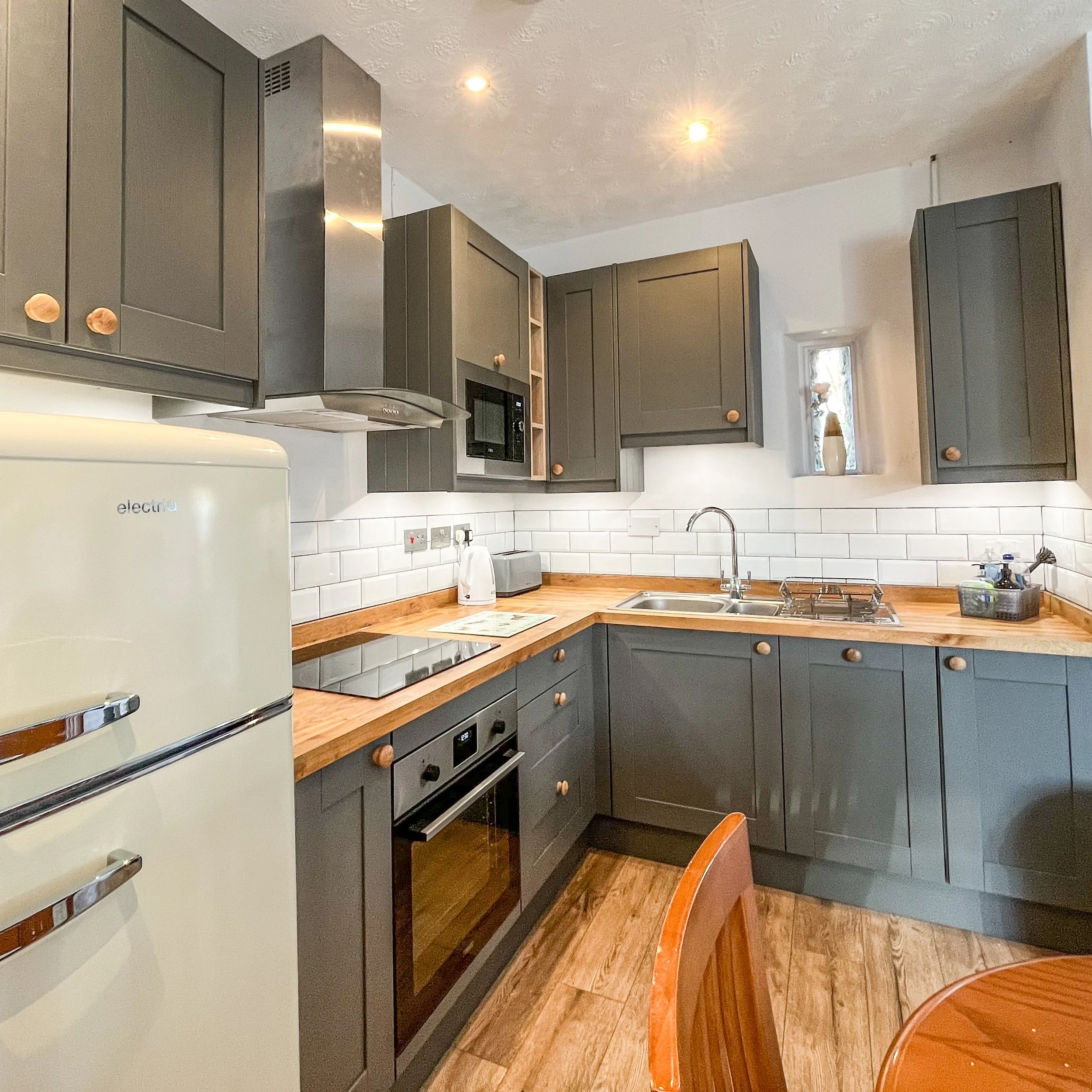A kitchen with gray cabinets, wooden countertops, white subway tile backsplash, and appliances including a refrigerator, microwave, and stovetop. There's a small window with a decorative vase and the floor is wood.