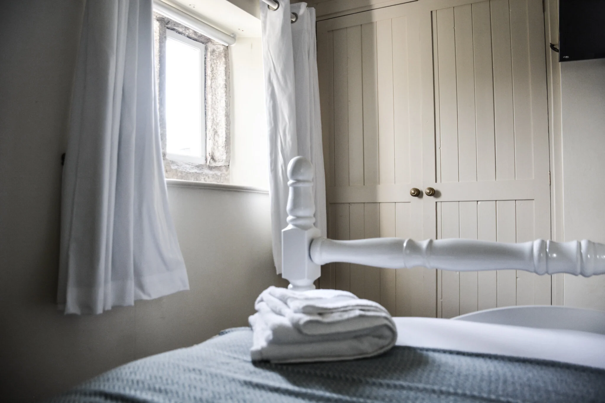 A bedroom corner with a bed, white linens, and a stack of folded towels on top. There is a window with white curtains and a stone frame, and a closed white wooden cabinet with brass knobs in the background.
