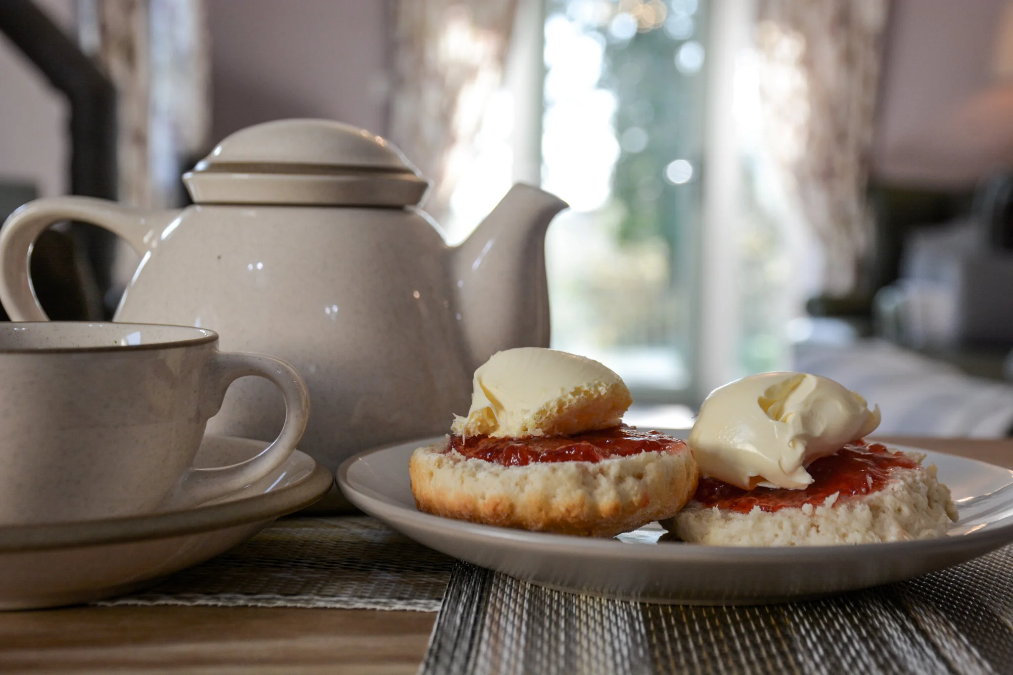 A cup and saucer, a teapot, and a plate with two scones topped with clotted cream and jam on a wooden table.