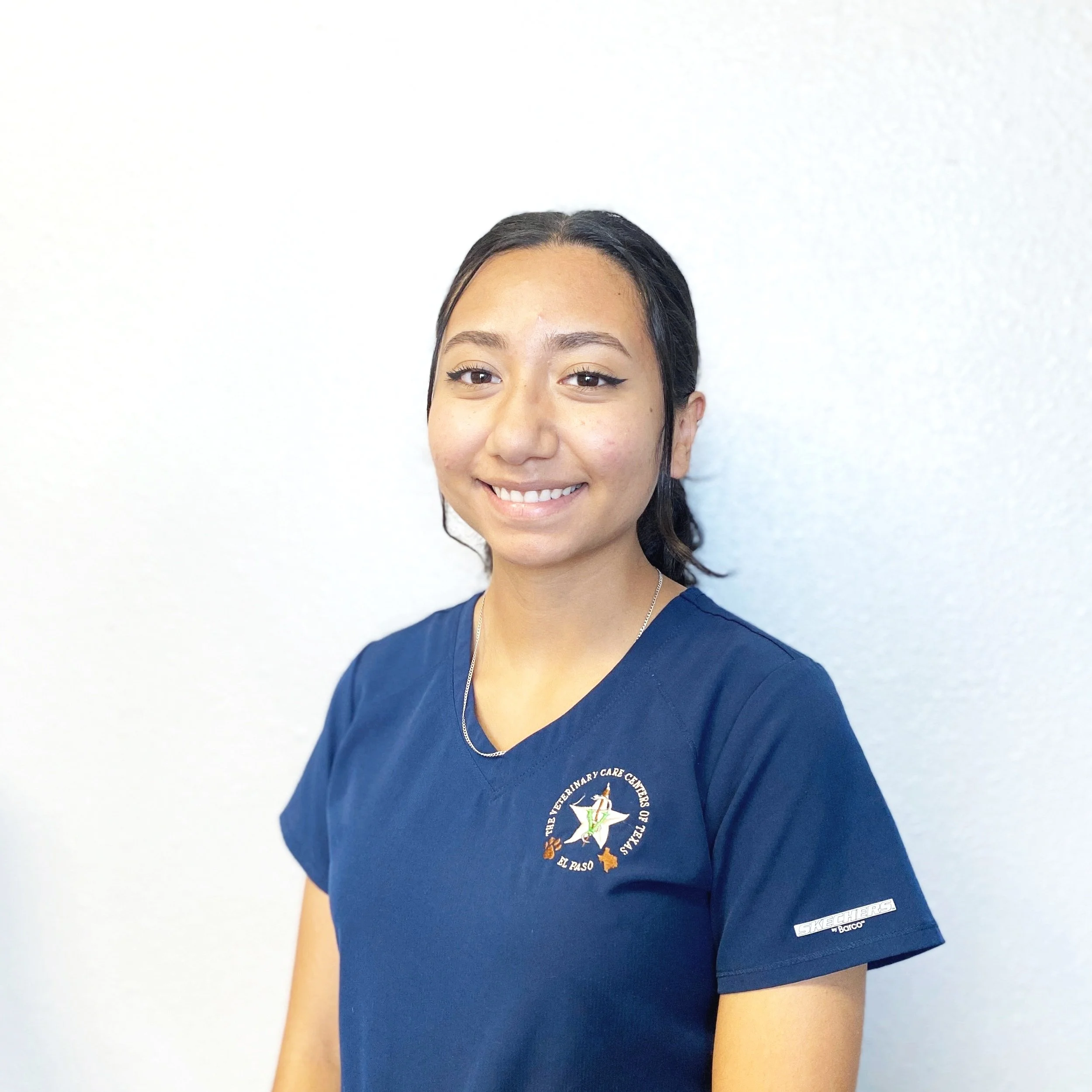 A woman wearing navy scrub shirt with a logo representing The Veterinary Care Centers of Texas, standing against a white wall, smiling at the camera.