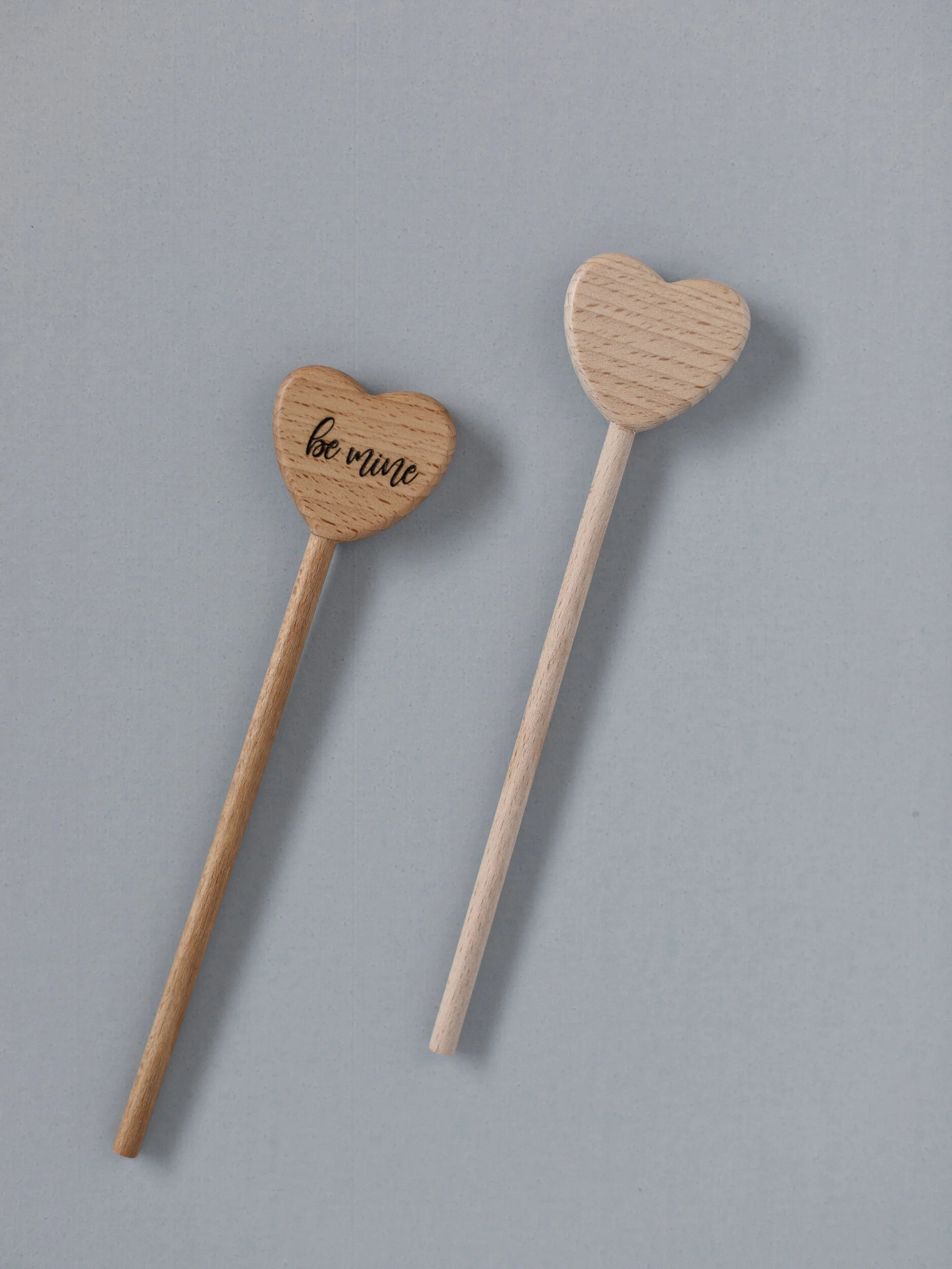 Two wooden heart-shaped spoons, one with 'be mine' written on it, on a light blue background.