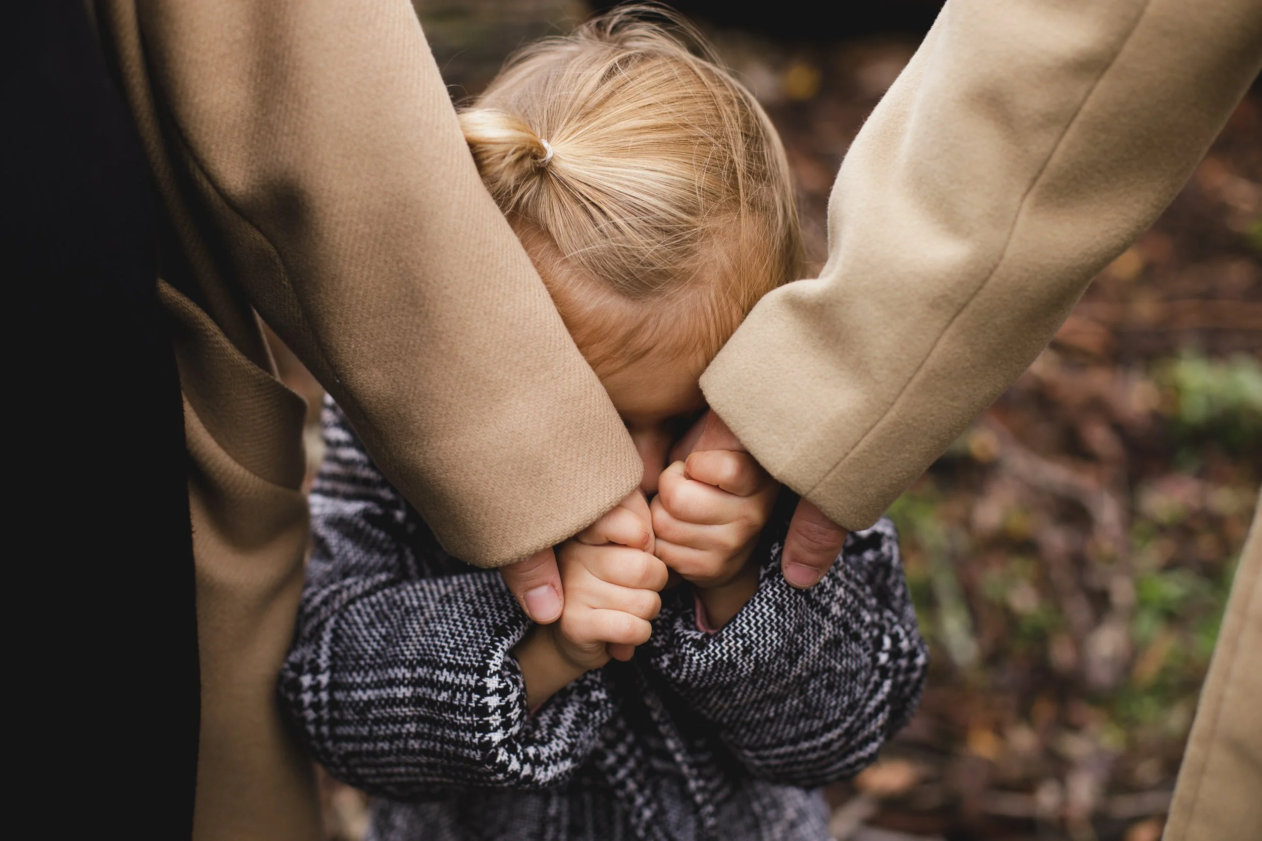 A young girl with blonde hair in a braid, wearing a gray and black sweater, being comforted by two adults in tan coats; one adult is holding her head and the other is holding her hand as she appears upset or crying outdoors.