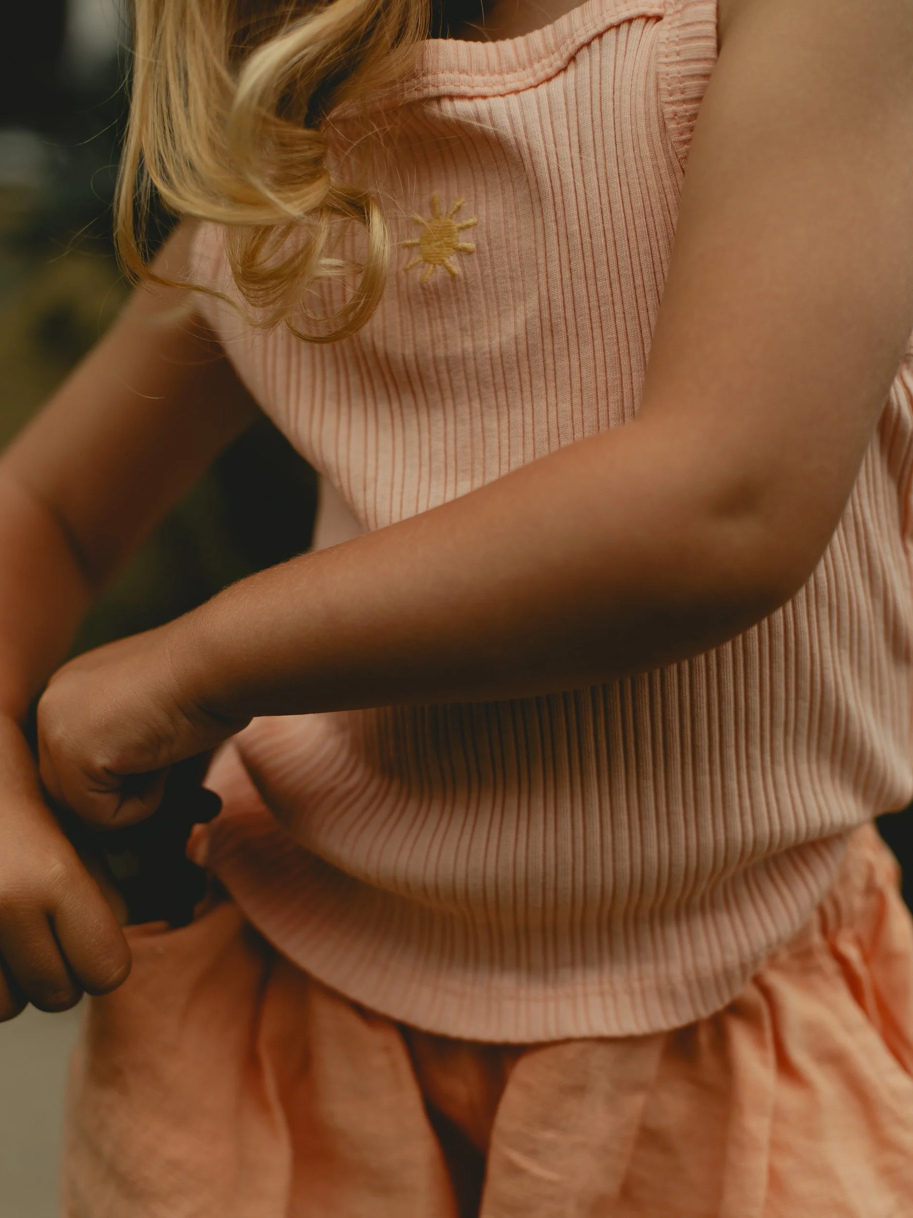 A young girl wearing a peach-colored sleeveless top with a yellow sun embroidered on it and matching peach shorts.