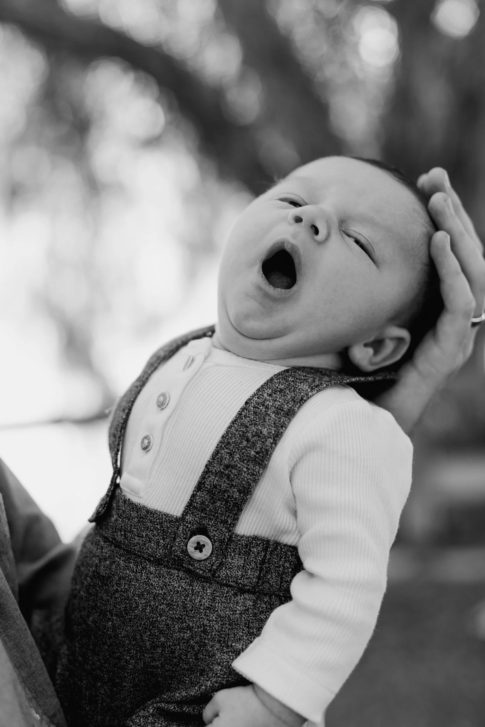 A baby yawning and being gently held by an adult's hand, outdoors with blurred trees in the background.