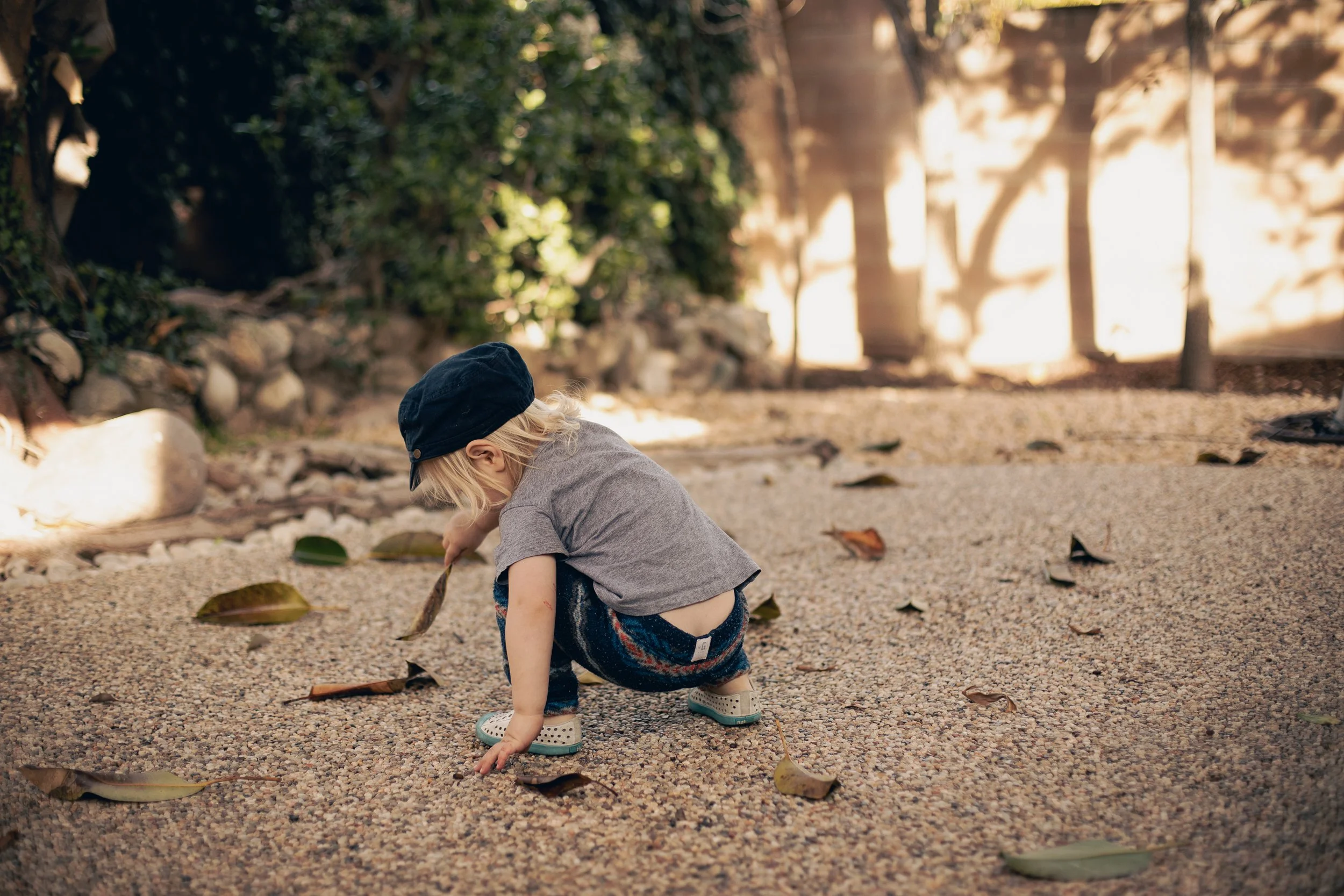 A young child crouching on a gravel pathway, picking up a fallen leaf. The child is wearing a dark cap, gray t-shirt, patterned shorts, and rubber shoes. There are scattered leaves around, with trees and a fence in the background.