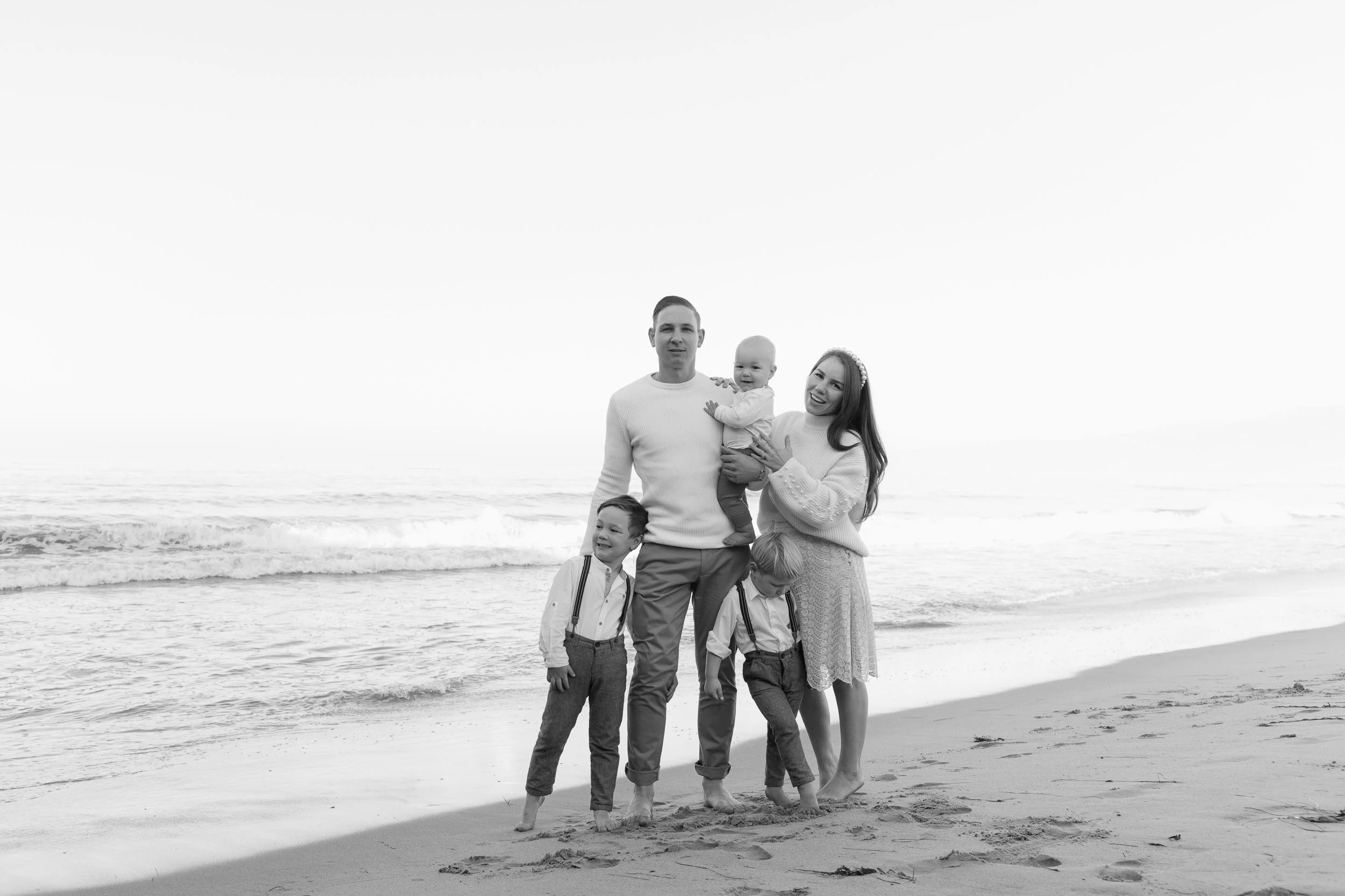Black and white photo of a happy family of five on the beach, with the ocean waves in the background. The mother and father are holding a baby, and two young boys are standing on the sand.