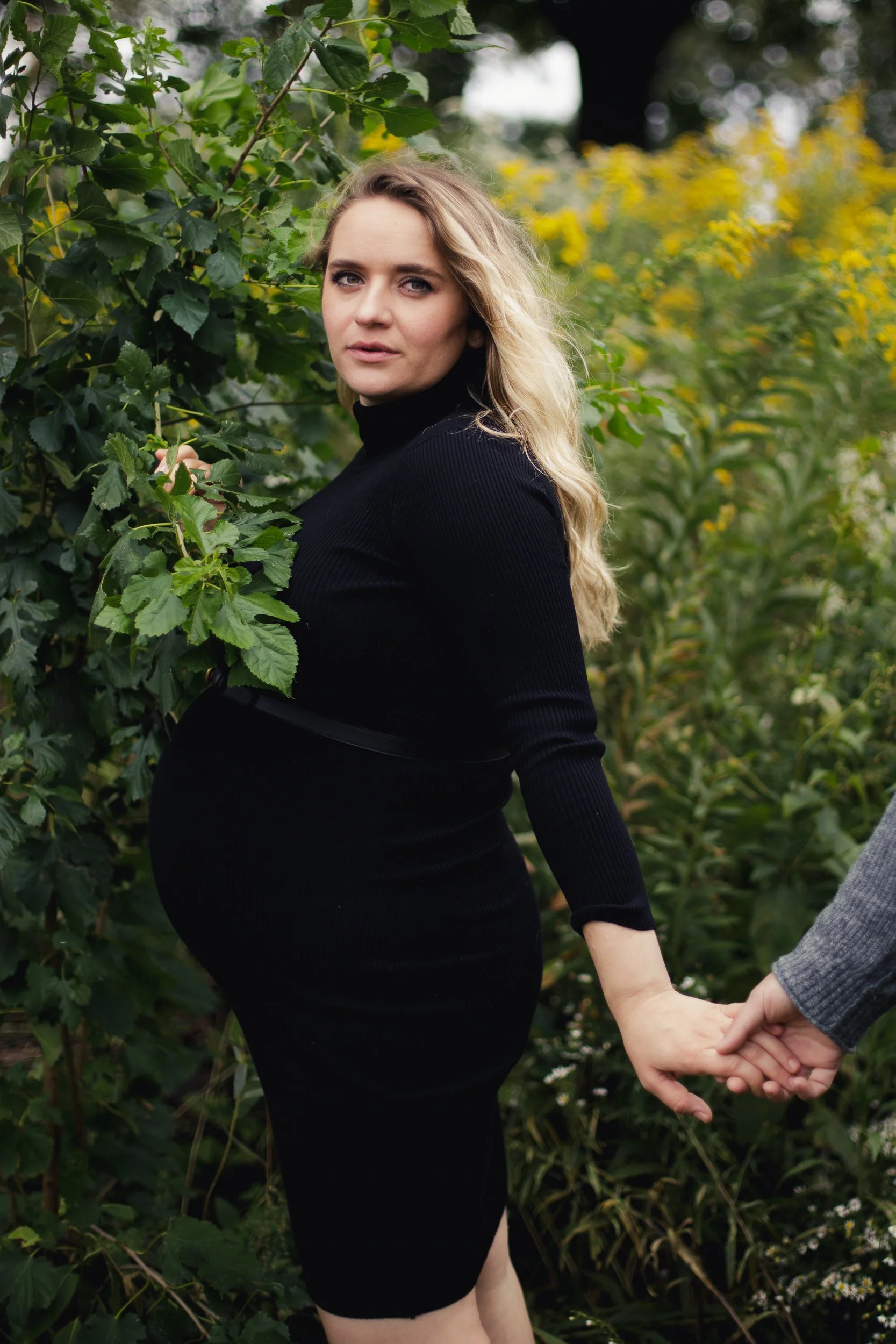 A pregnant woman in a black dress standing outdoors, holding hands with a person in a gray suit, surrounded by green foliage and yellow flowers.