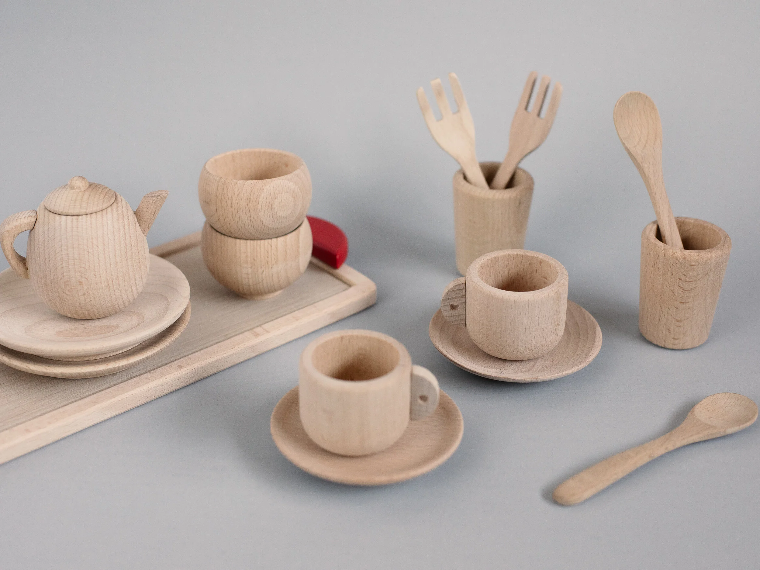 Set of miniature wooden toy tea set with teapot, cups on saucers, and utensils on a light gray surface.