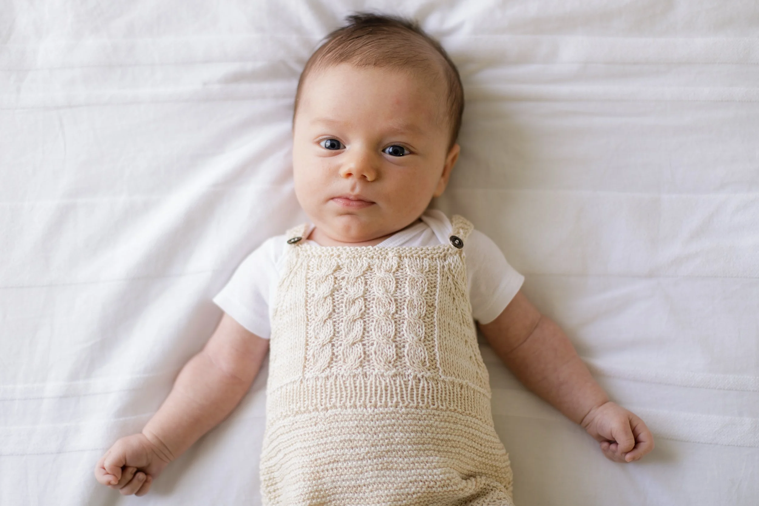 A baby lying on a white bed, wearing cream-colored knitted overalls and a white T-shirt, looking at the camera with a neutral expression.