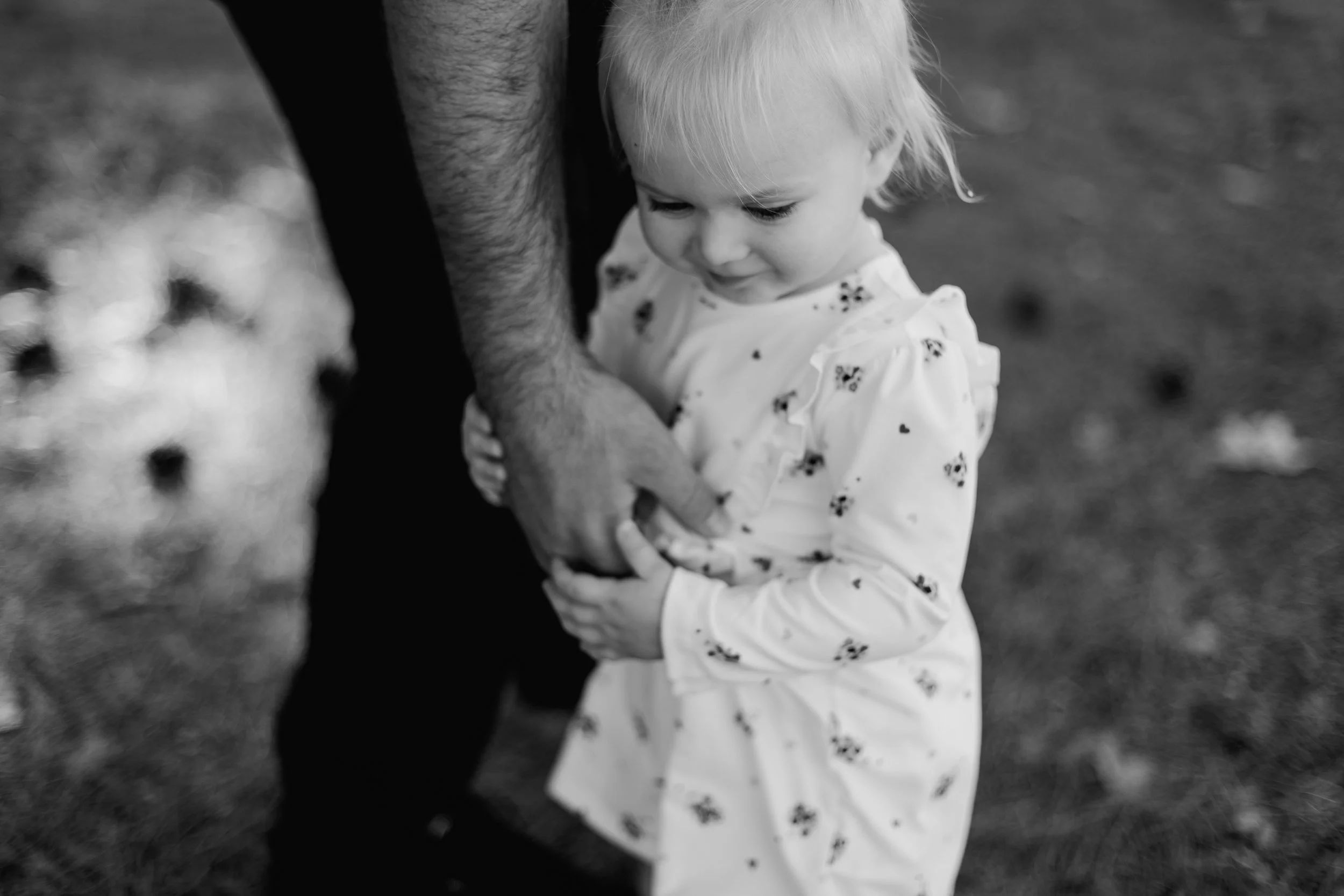 A young girl in a floral dress holding onto an adult's arm outdoors, with the focus on her face and hands, black and white photo.