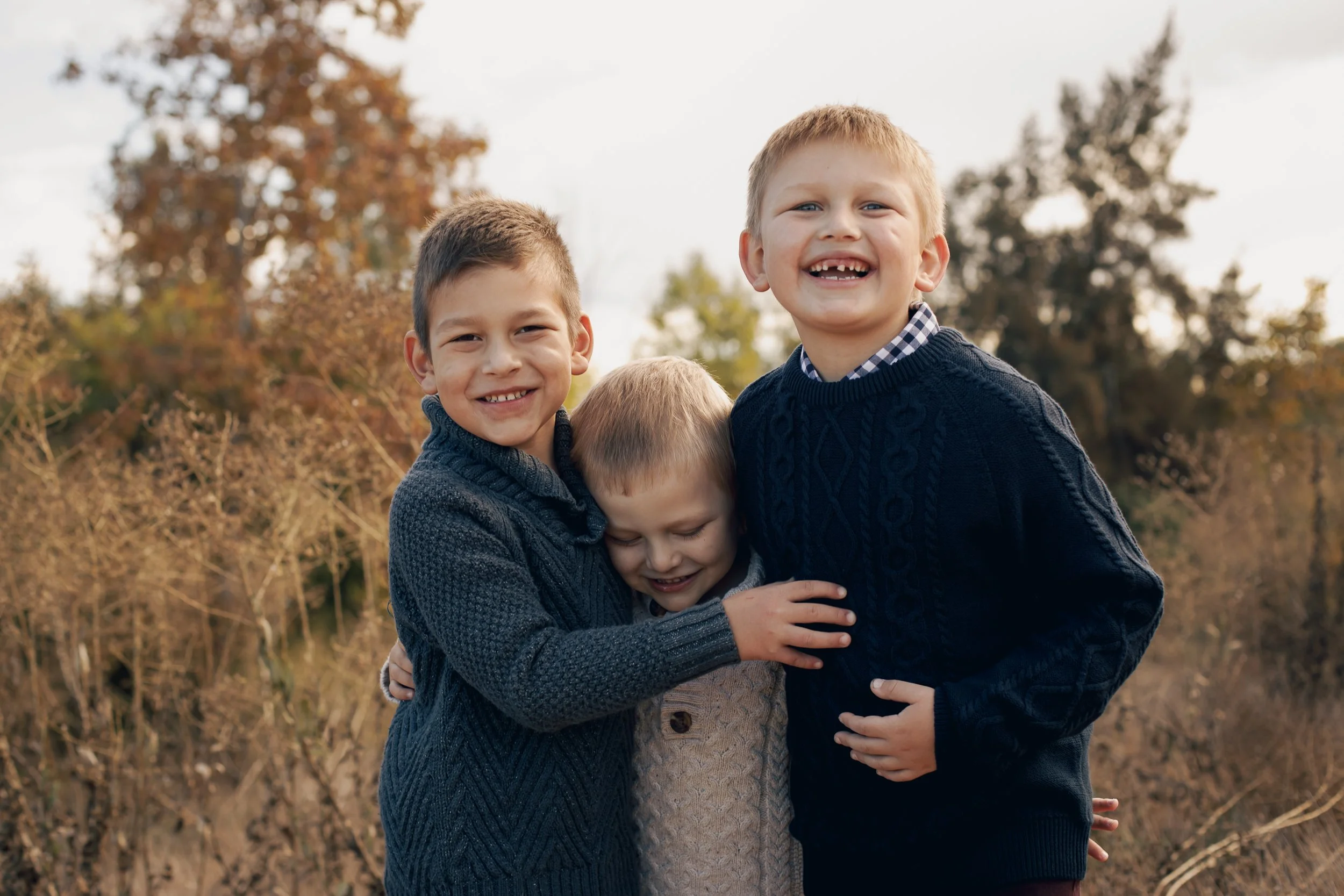 Three young boys outdoors, smiling and hugging each other, surrounded by trees with fall-colored leaves.