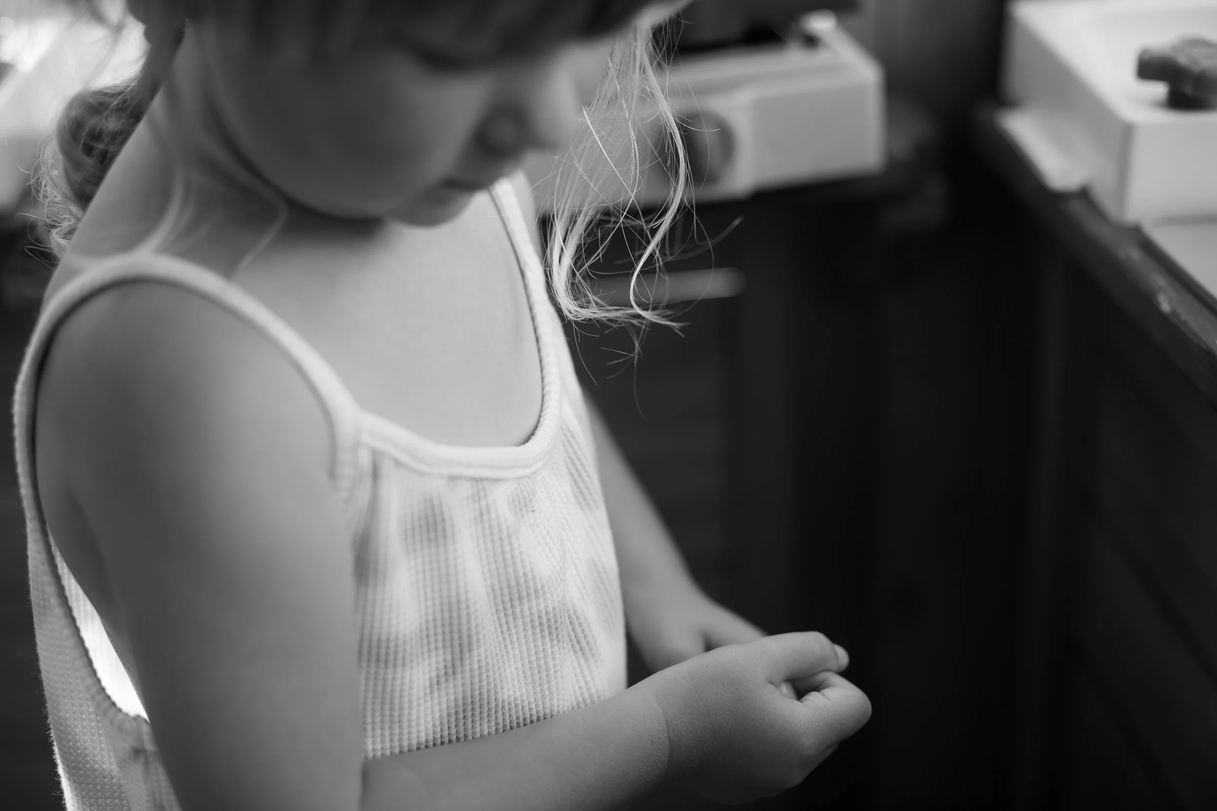 A young girl with long hair looking down in a kitchen or workshop area, wearing a sleeveless top.