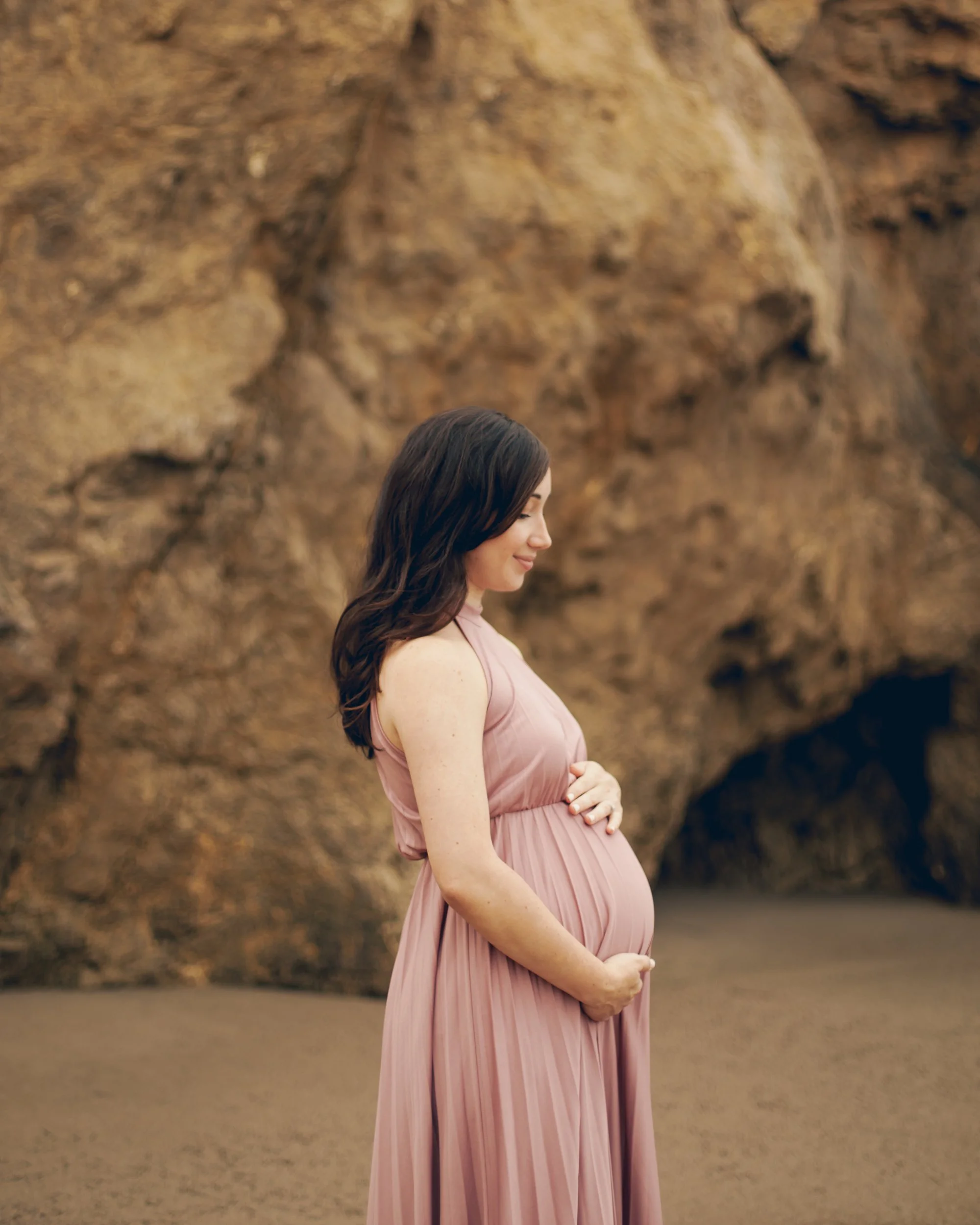A pregnant woman in a pink dress standing on a beach with rocky formations in the background.
