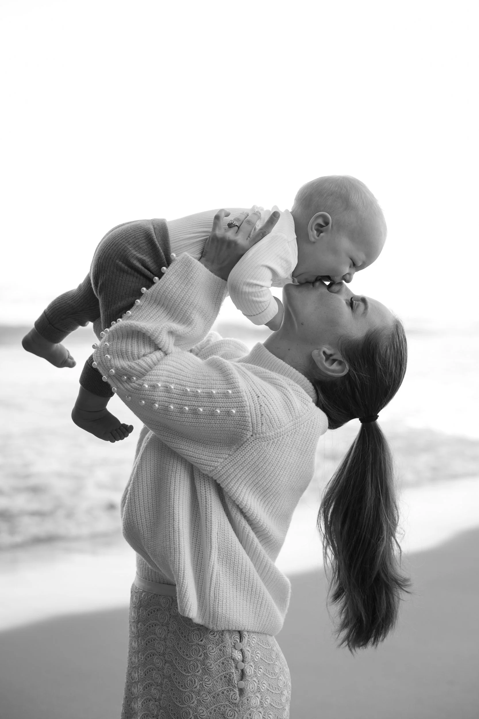 A woman holding a smiling baby boy near her face on a beach at sunset or sunrise, with the ocean in the background, in black and white.