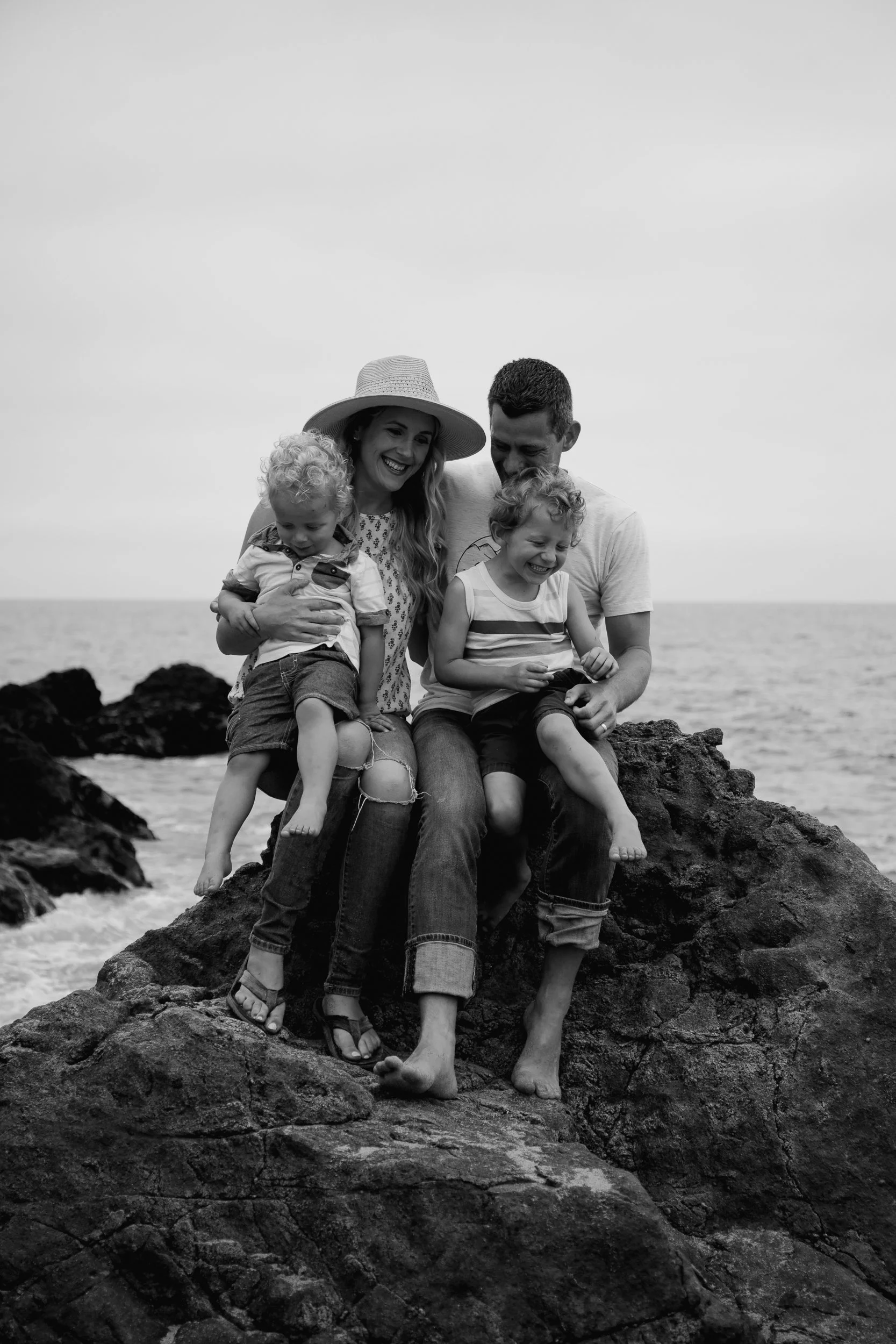 A family of four sitting on rocks by the ocean, laughing and enjoying a day at the beach. The mother is wearing a wide-brimmed hat, and the children are barefoot.