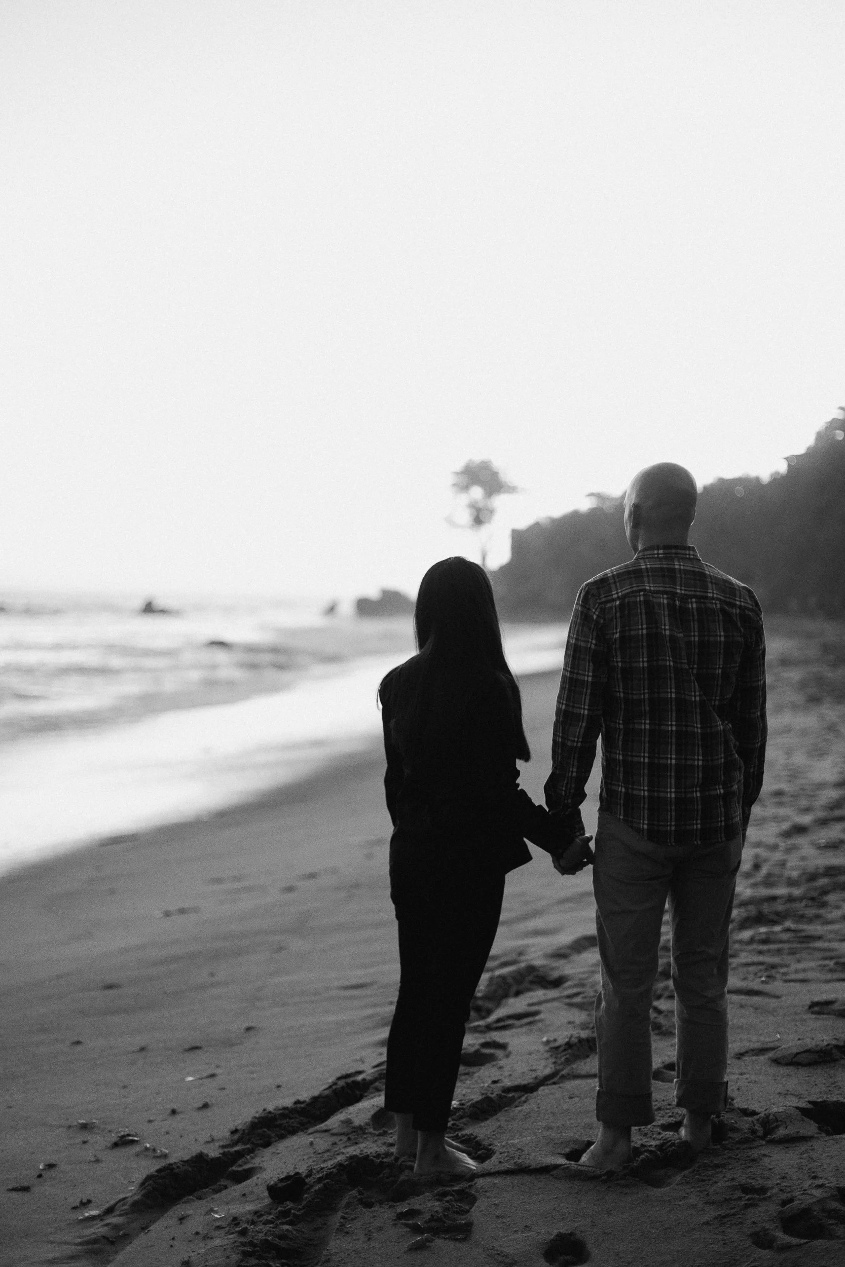 A black and white photo of a couple holding hands and walking on the beach at sunset, facing the ocean.