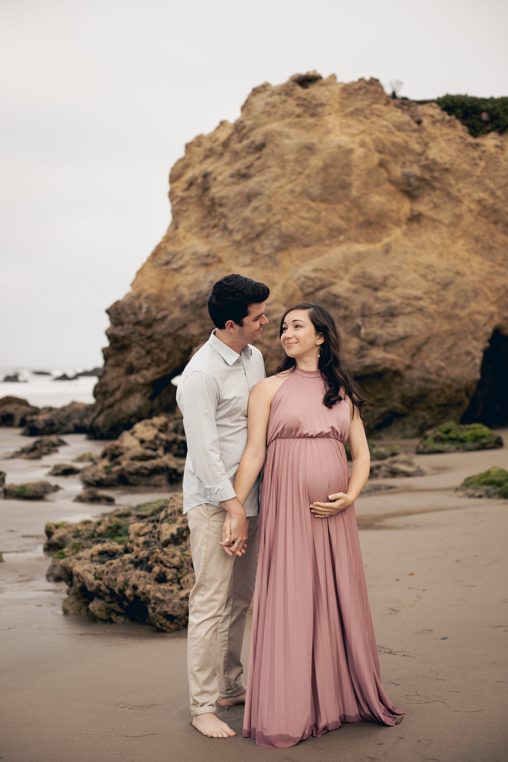 A pregnant woman in a pink pleated dress and a man in a light-colored shirt and pants holding hands on a beach with large rocks.