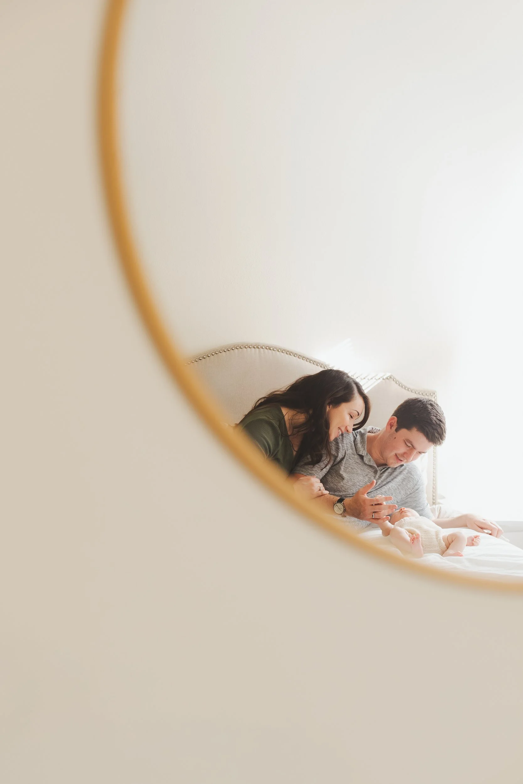 A couple smiling and looking at their baby through a mirror, with the mirror partial view showing the family on a bed.