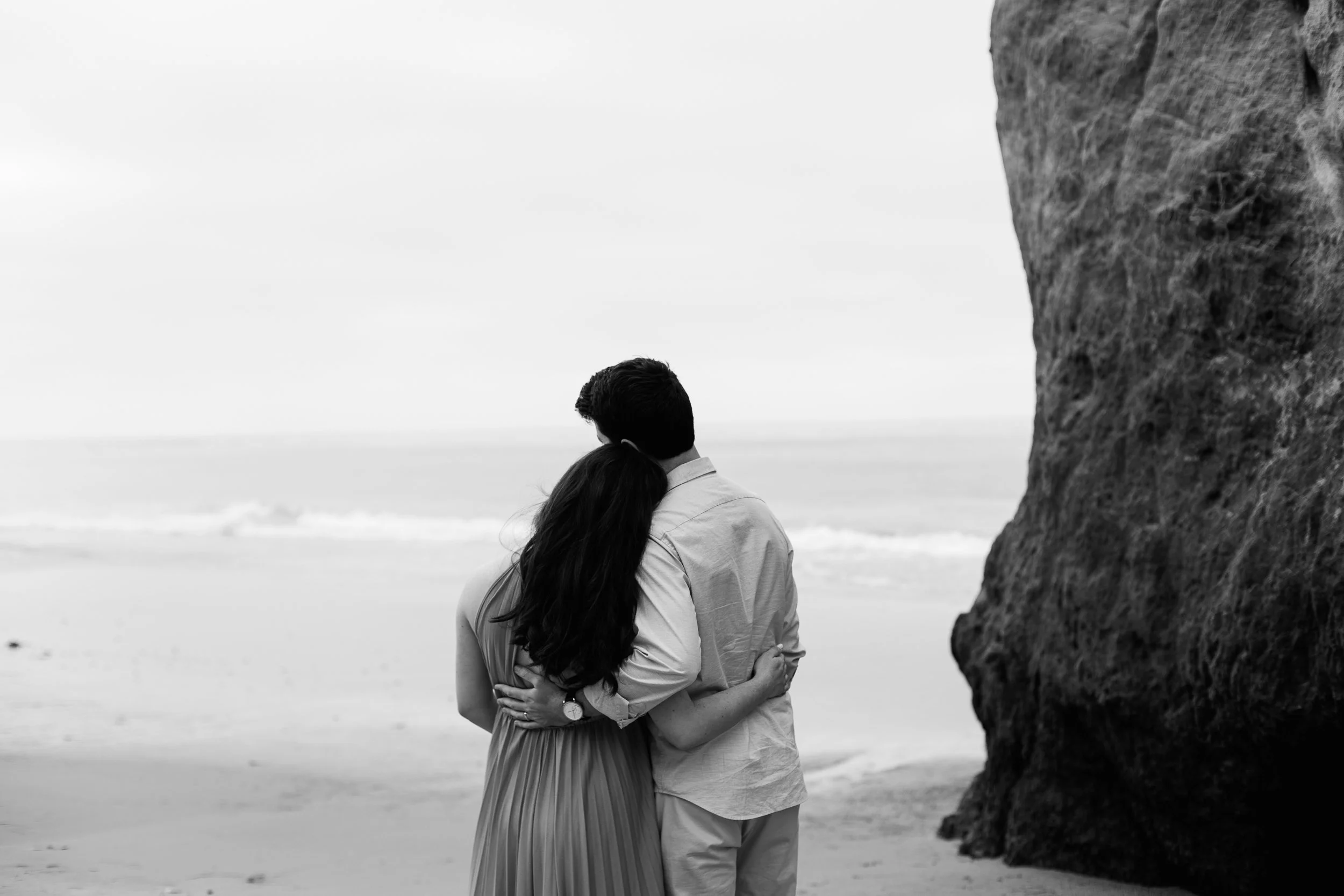 A black and white photo of a couple standing on a beach, hugging and looking out at the ocean, with a large rock formation on the right side.