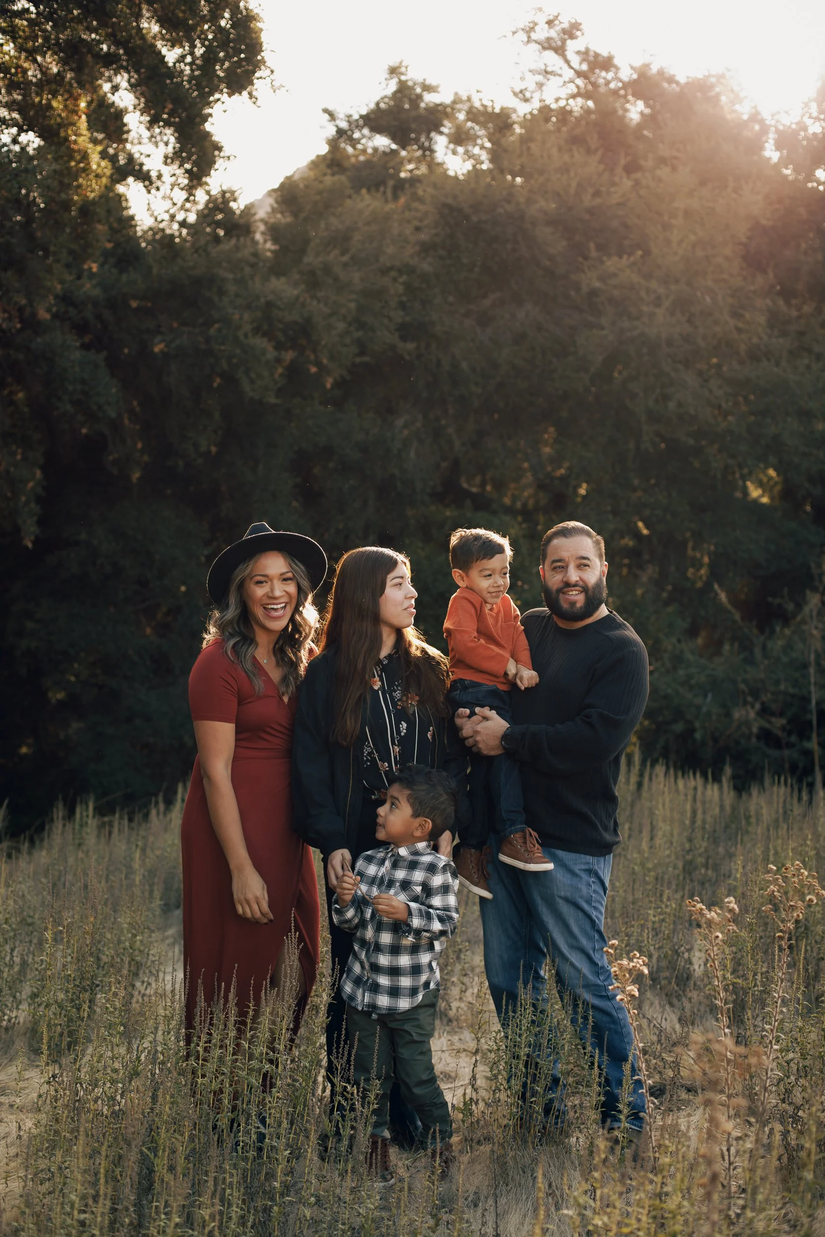 Family of five standing outdoors in a field with trees in the background, smiling and enjoying the sunny day.