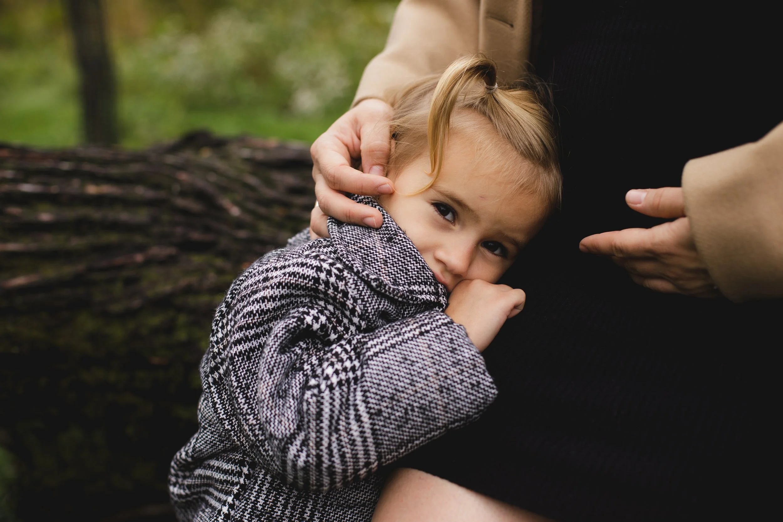 A young girl with light brown hair and a gray patterned coat is hugging a pregnant woman's belly while resting her head on it. The background shows a blurred outdoor setting with green grass and trees.