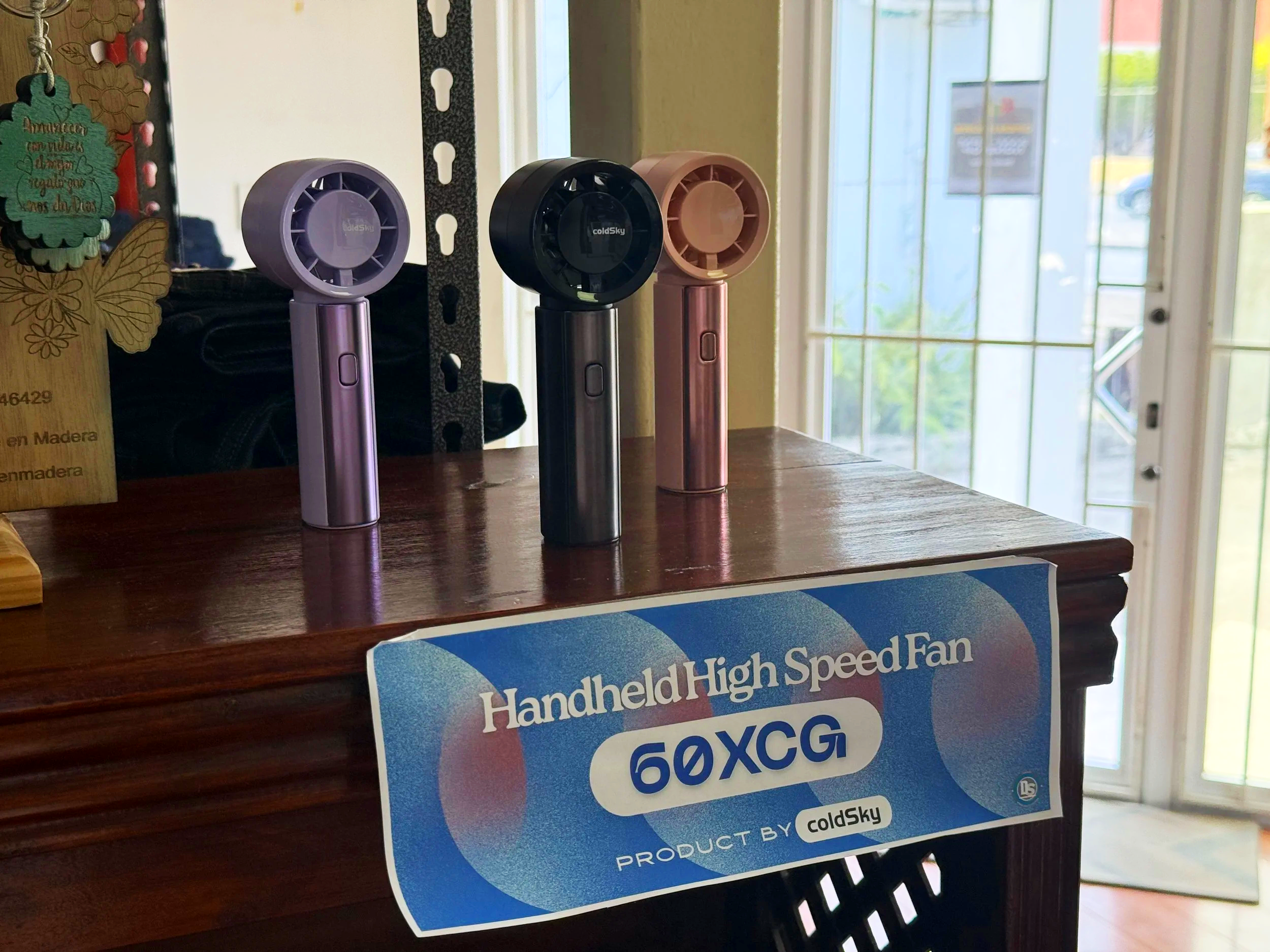 Three handheld high speed fans in purple, black, and pink colors are displayed on a wooden surface near a window, along with a blue promotional sign.