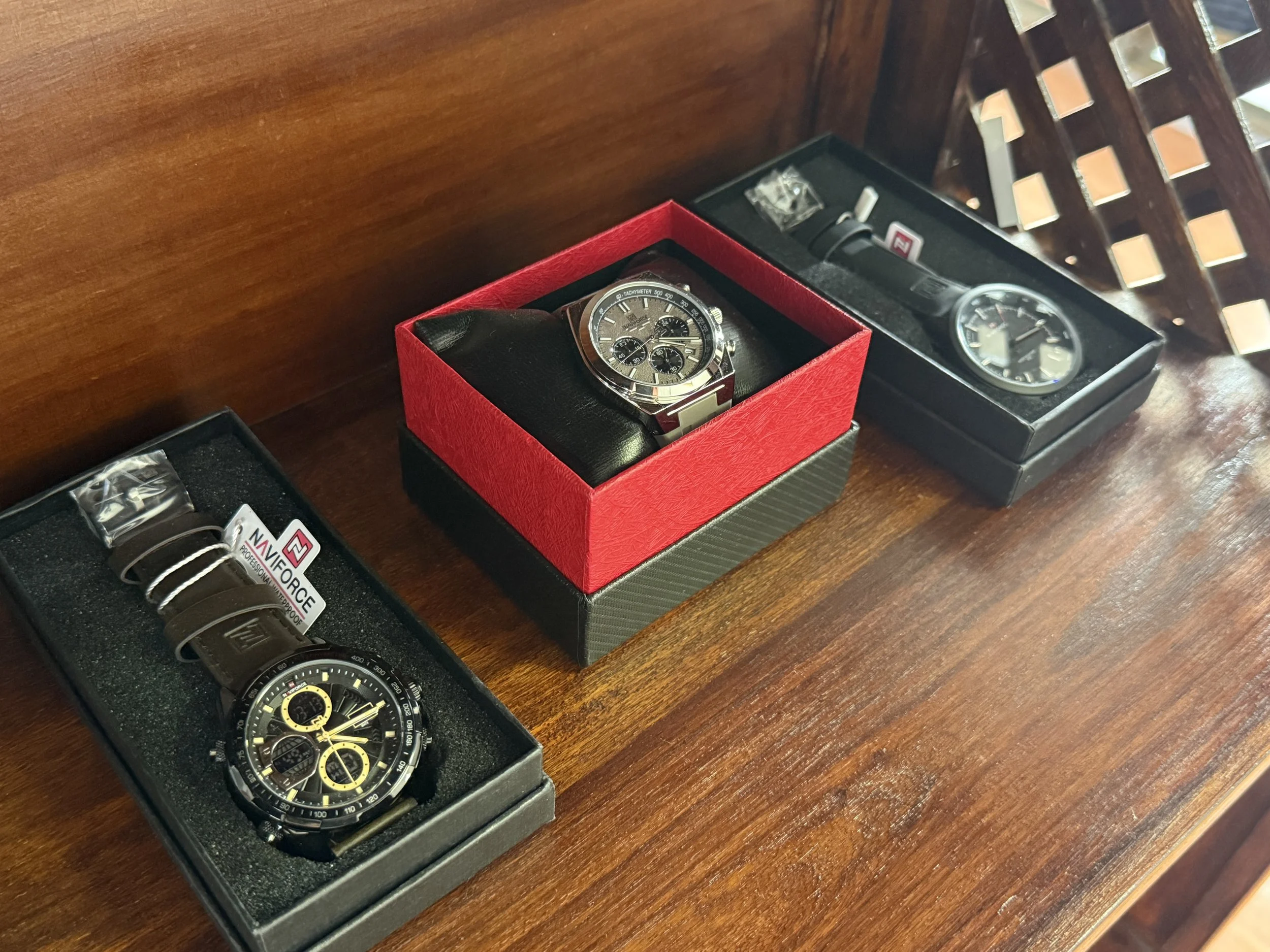 Three watches in boxes on a wooden surface, with a brown watch in a red box, a black watch in a black box, and a black and yellow watch in a black box.