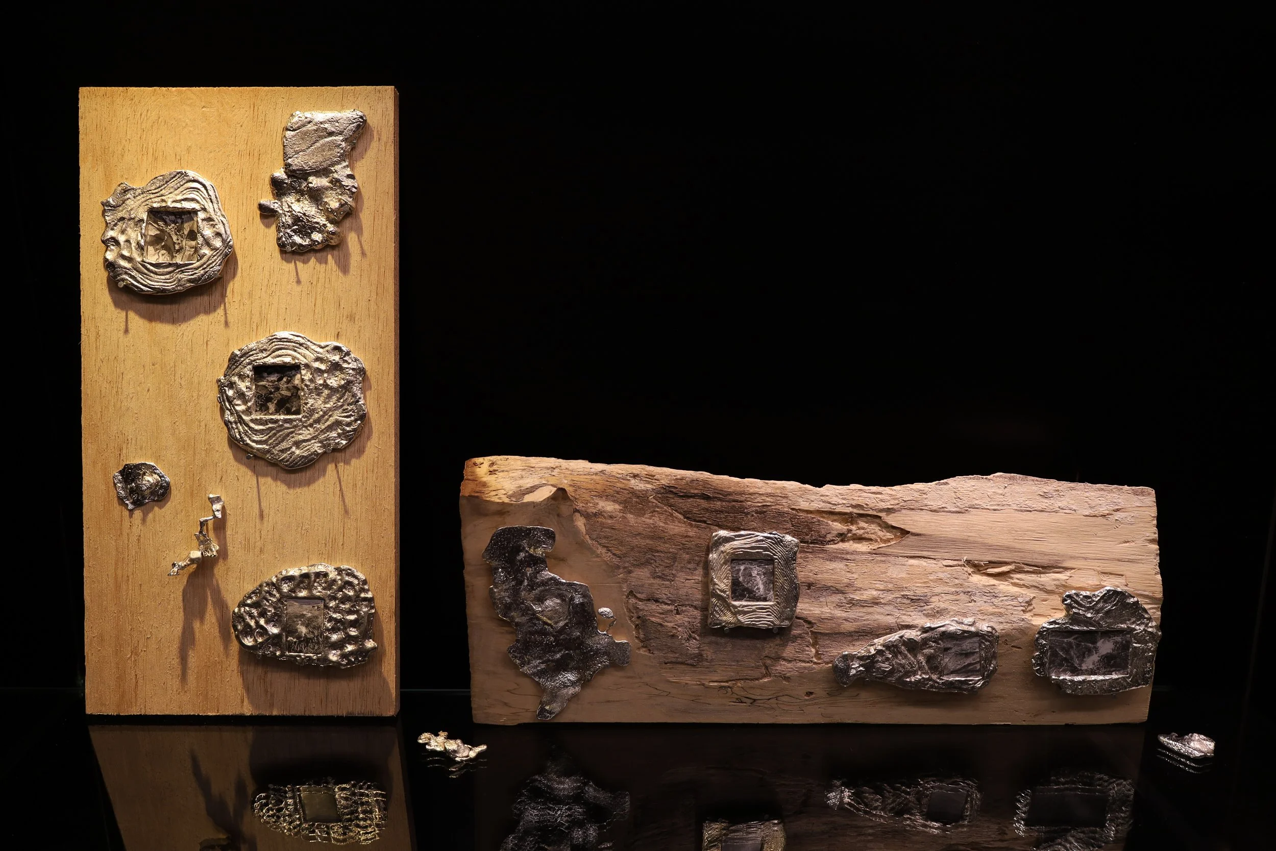 Vertical plank of wood & horizontal weathered wood block with many organic shaped pewter pieces with textured wave-like ridges. Some of the pieces are picture frames, housing small photos of ocean waves and rocky cliffs.