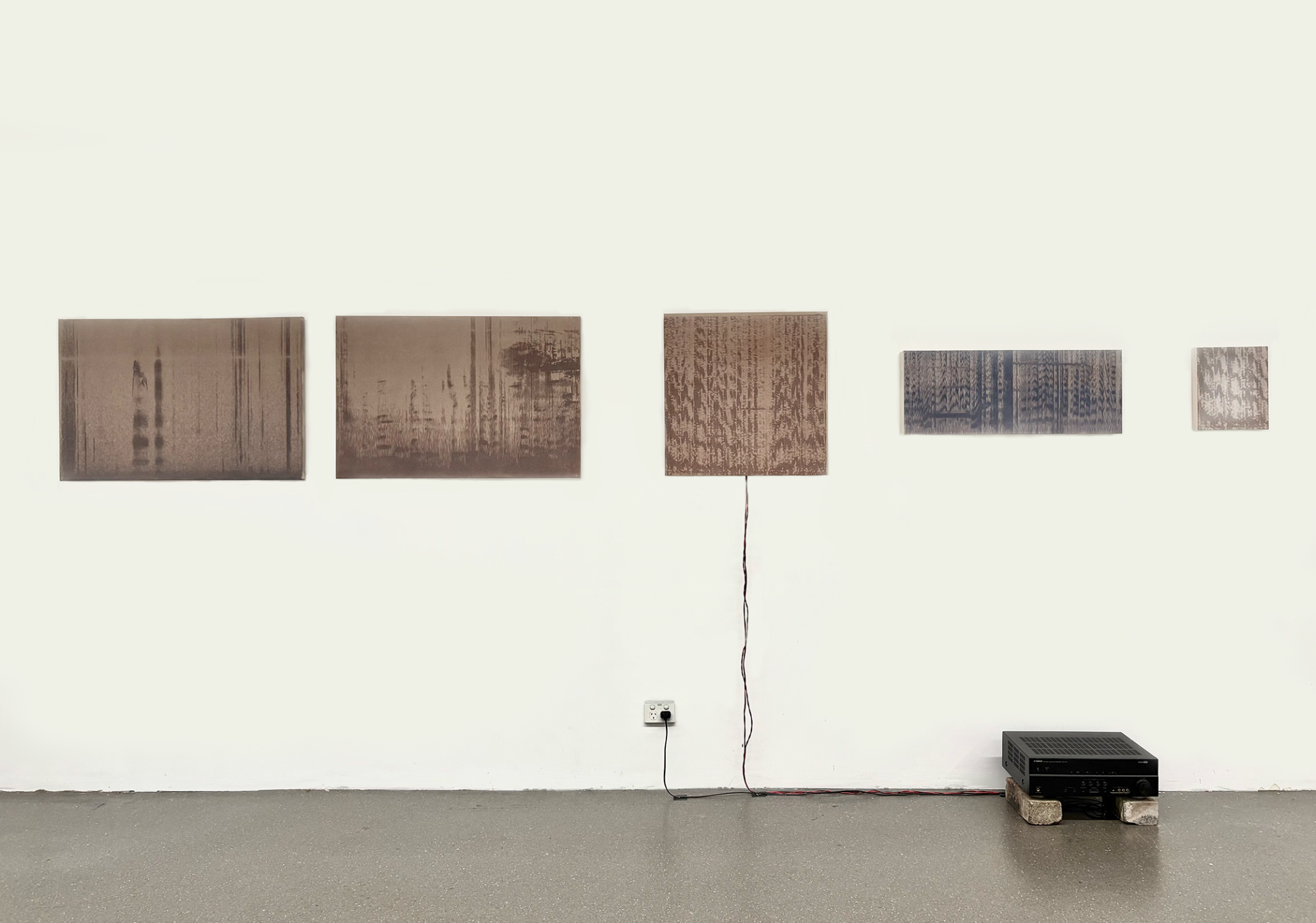 Noah Mair, Armoured Letter, 2026. 5 brown, dyed canvases with sound wave patterns installed on a wall. A wire extends down from the middle canvas, connecting to a speaker system on brick stilts.