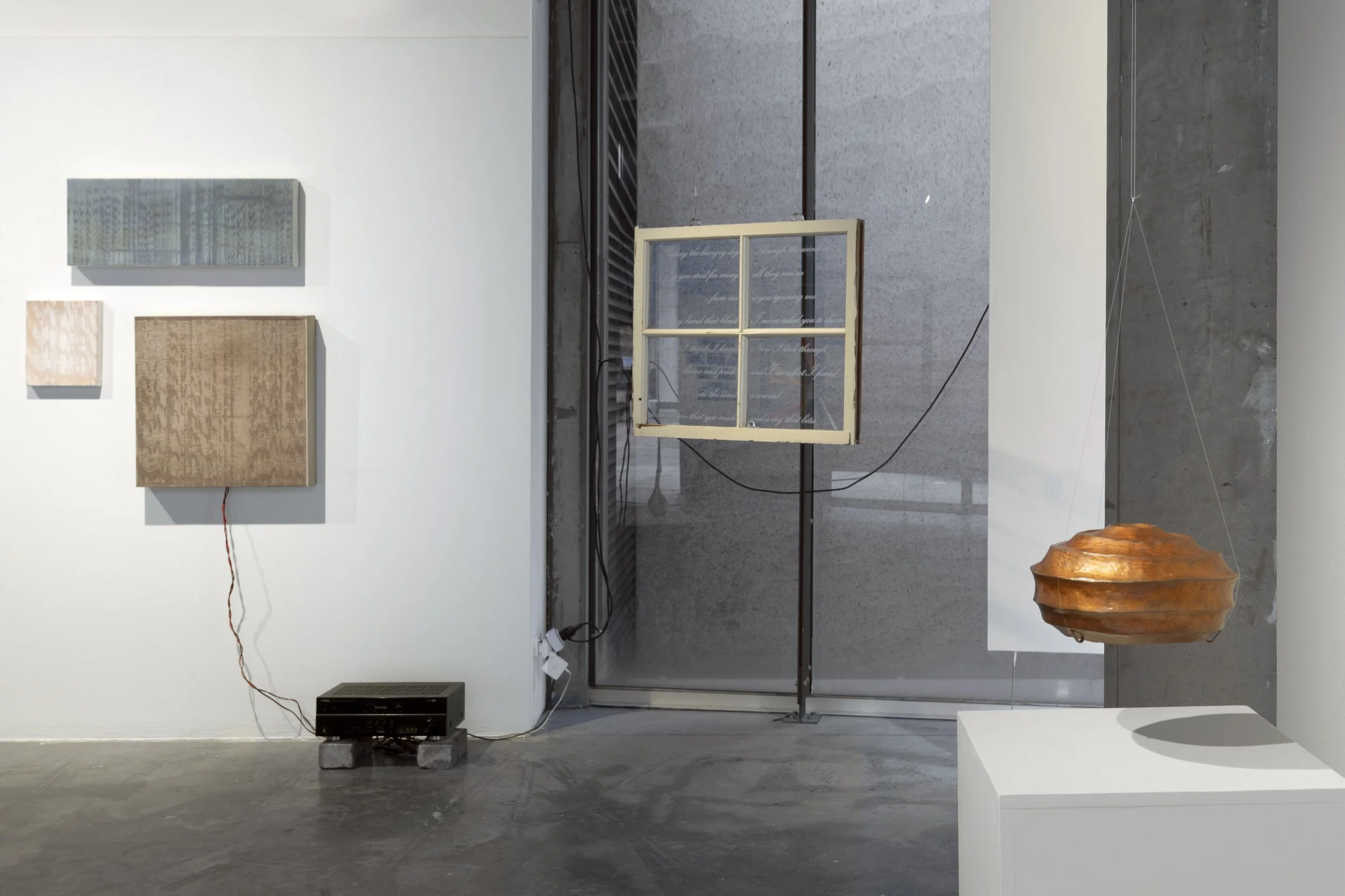 Left to right: 3 sculptures of dyed, patterened fabric in blue, beige and brown on a wall, connected to a speaker on the floor. Suspended window with white frame and text engraving. Suspended round copper sculpture.