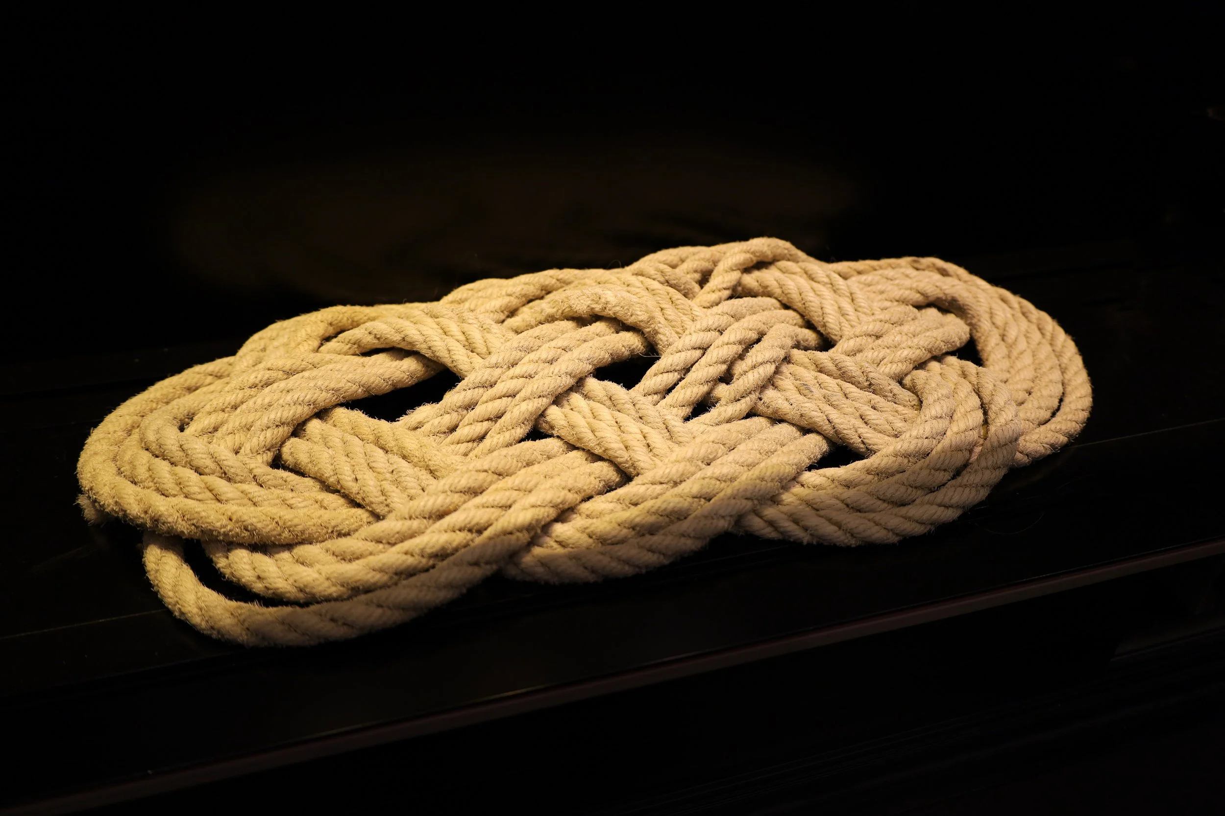 Large rope knot consisting of a braided pattern where each section is 3 rope widths thick. The rope is made of twisted brown fibers with a rough texture. The knot sits on a black background.