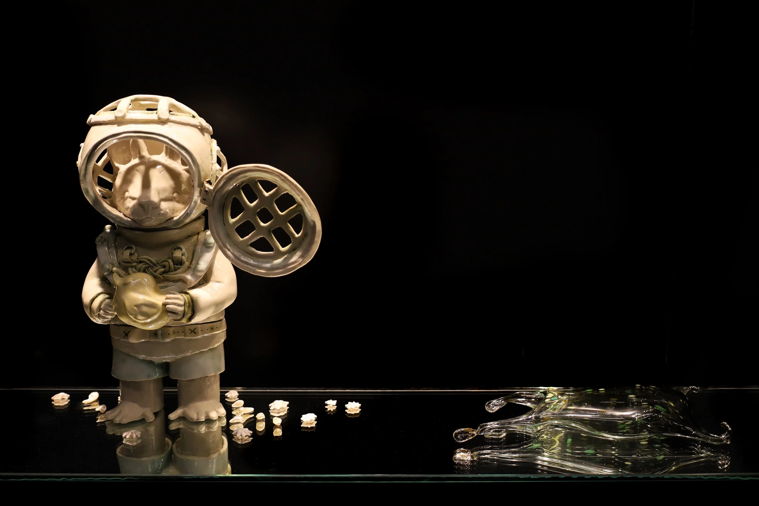 Left, ceramic sculpture of standing bat-like creature in an old diving suit with helmet shield open, holding a small duck, with shells scattered by its feet. Right, 2 organic shaped blown glass pieces in black and green.