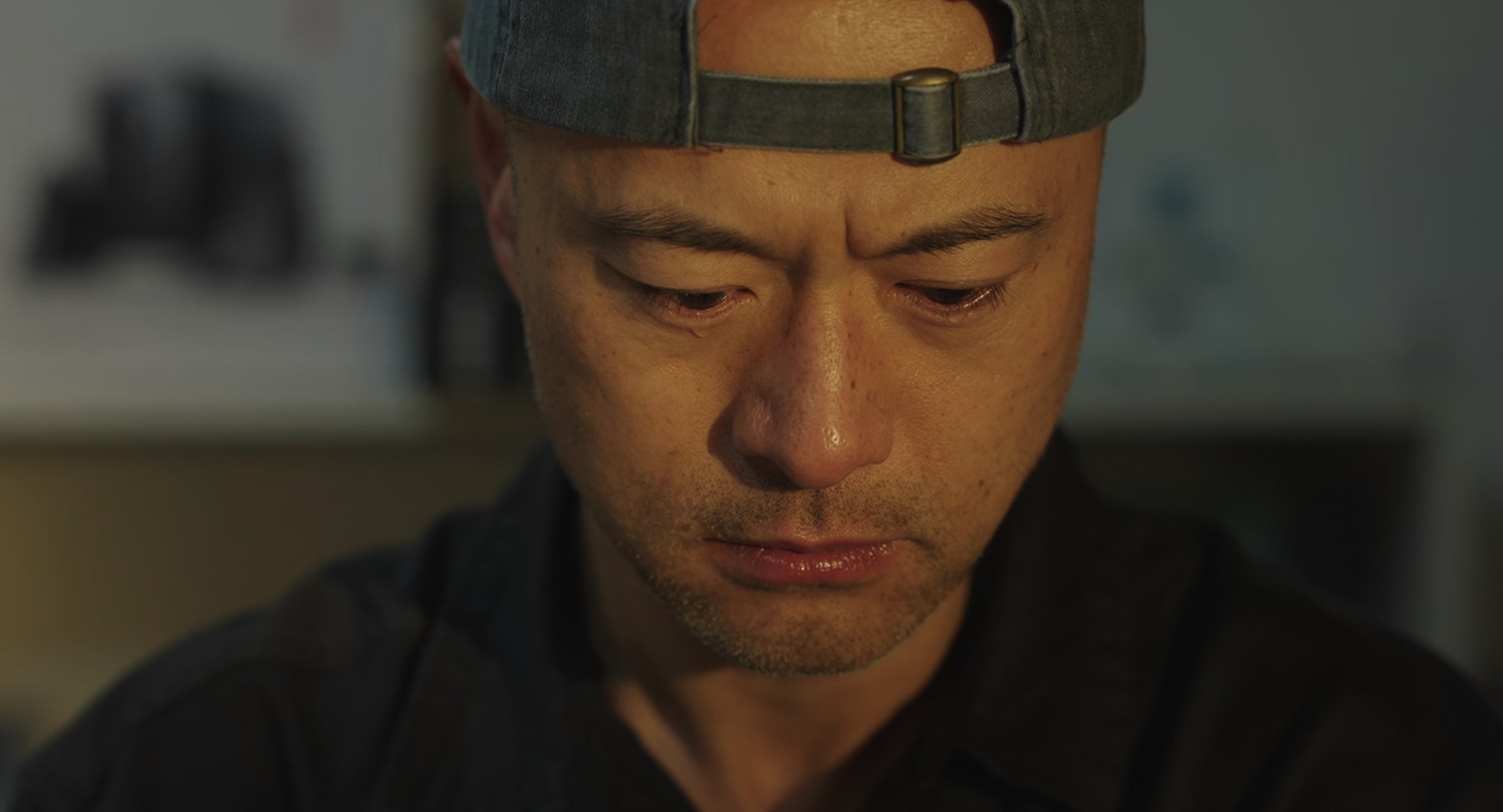 Close up image with warm lighting of a man wearing a cap looking downward with a serious expression.