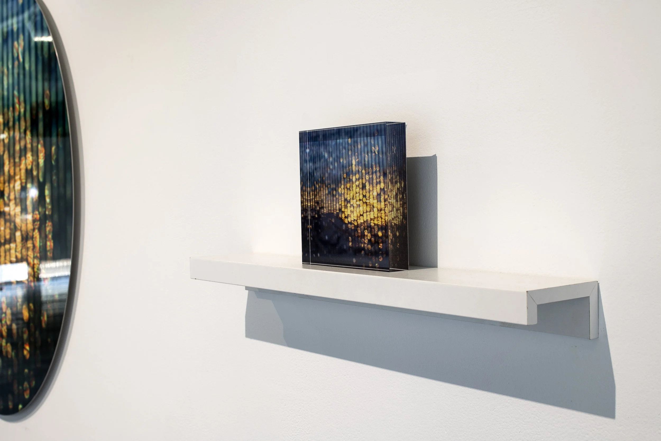 Small square block with printed image of lines of blurred, colour dots, from blue to yellow to black, on a white shelf.