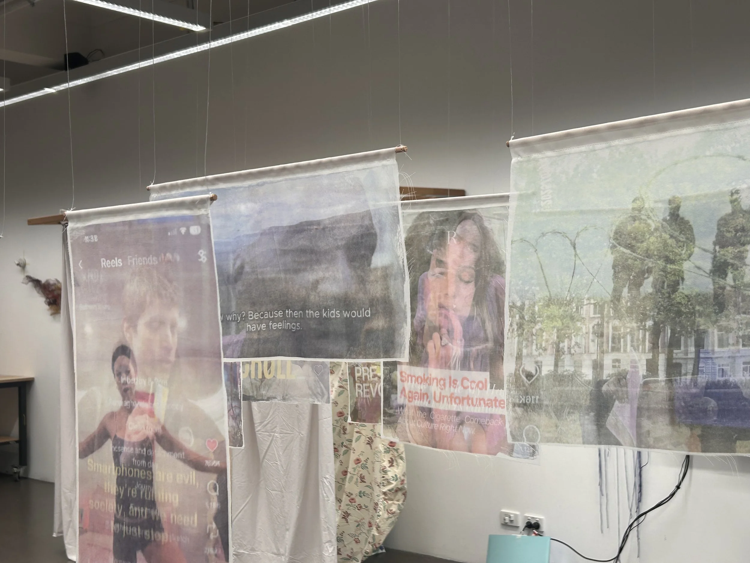 Amy Bailey, indebted, 2026. Many textiles on rods suspended from the ceiling with printed designs from Instagram reels, with some text reading: "Smoking Is cool Again, Unfortunately.".