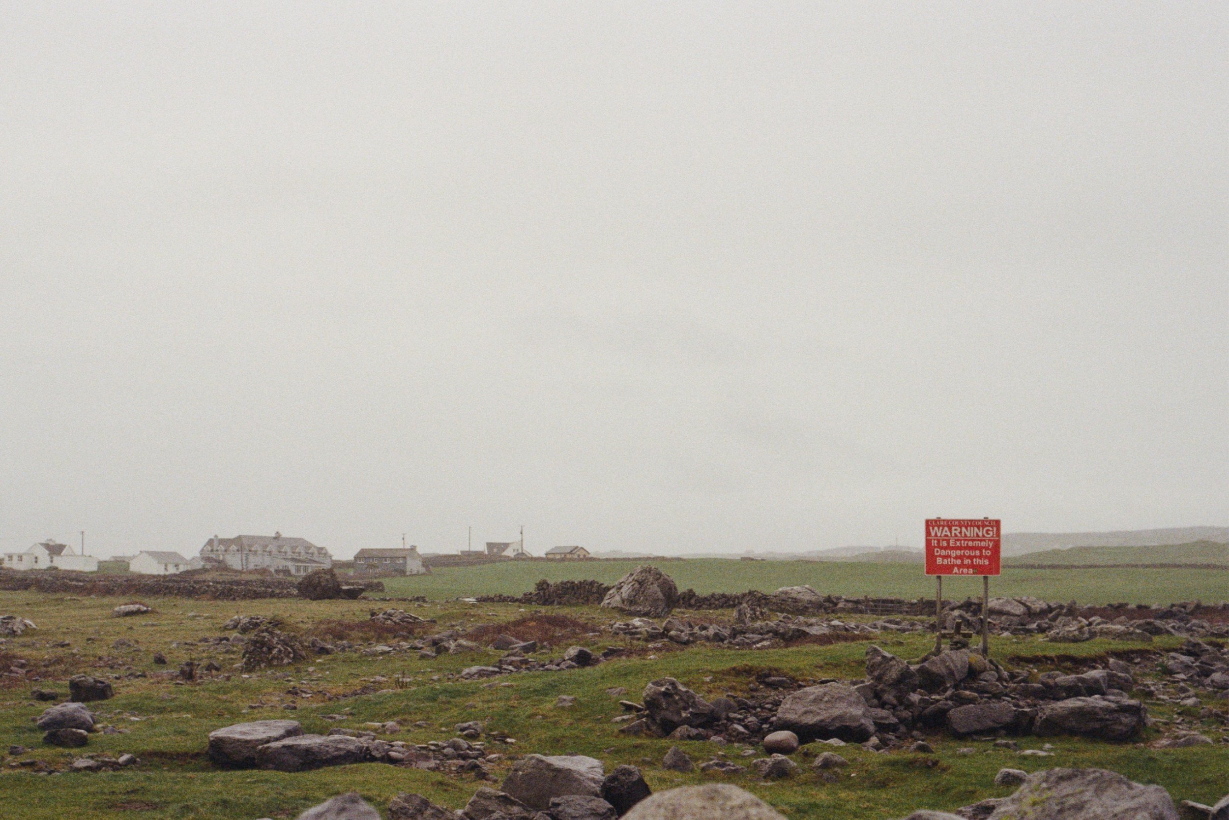 Charlie Boobar, Follow the signs, 2026. Photograph of a field of green grass and rocks with some buildings in the distance, and a red warning sign. The weather is grey and foggy.
