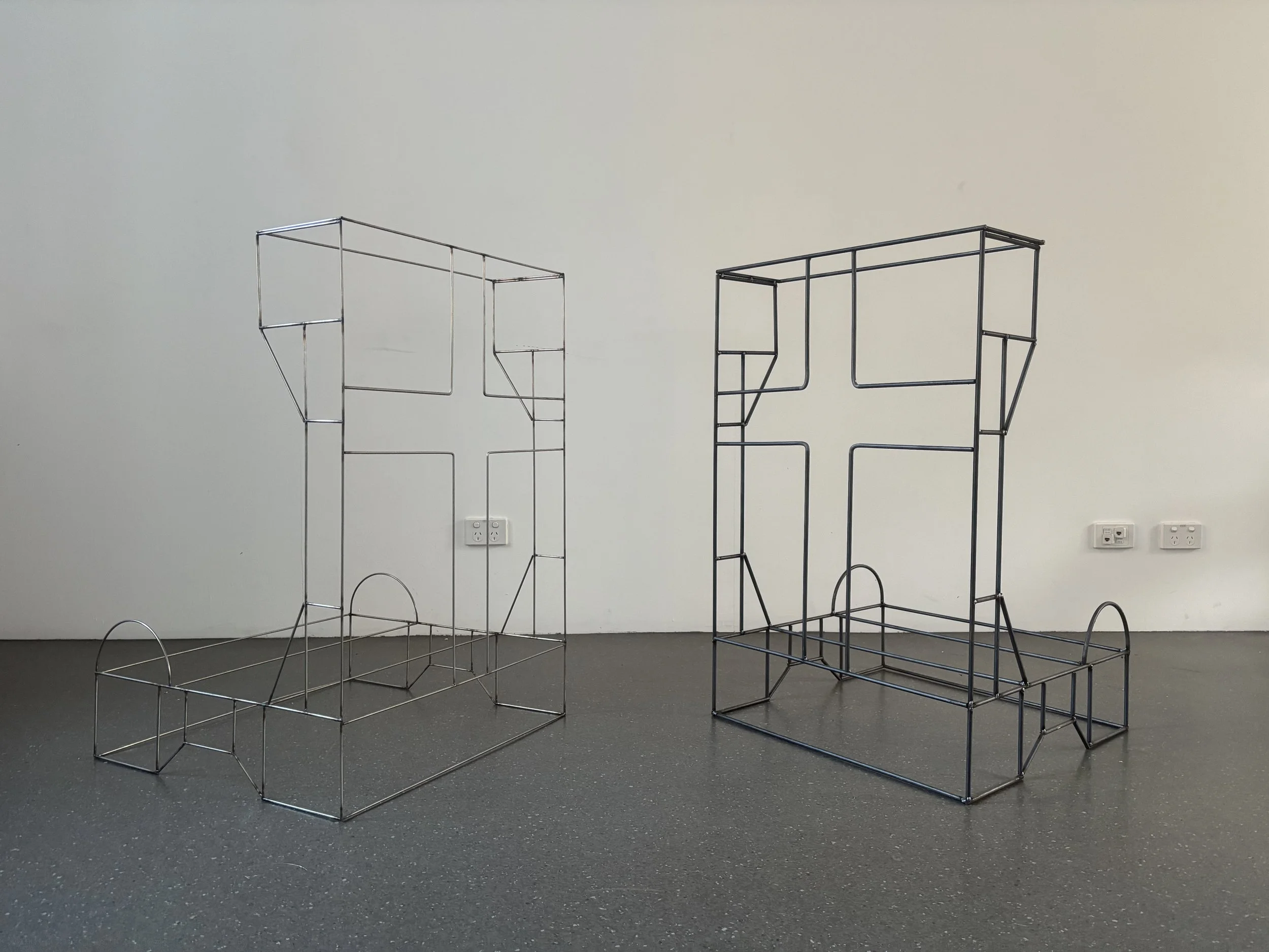 Ginia Caldwell, This Is Where I [Should} Pray, 2026. 2 large geometric sculptures of prayer or confession booths with cross designs. The sculptures are made of thin metal beams in light metallic and dark grey.