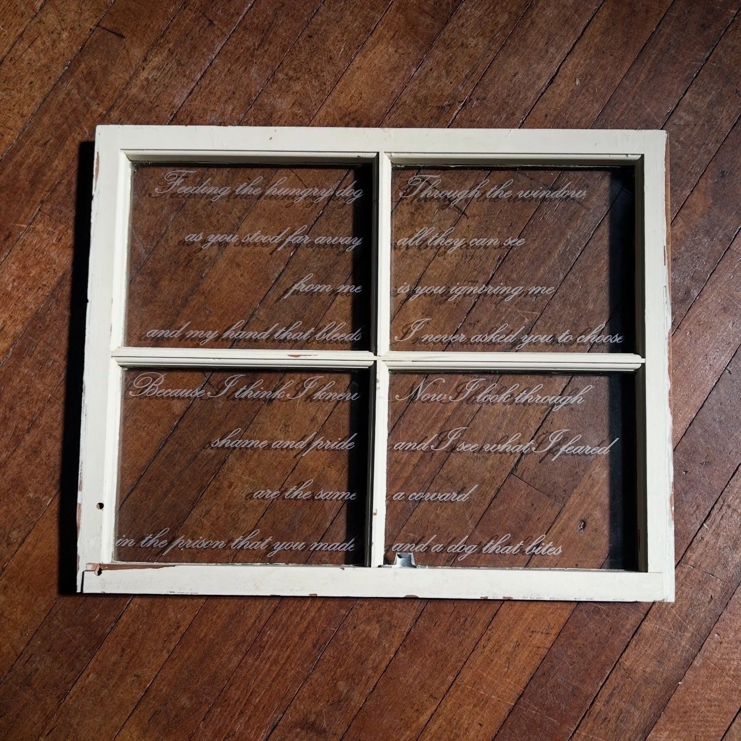 Edwina Darling, My Wound Is My Hope, 2026. A white framed window with textured cursive script on each glass pane. One pane reads: "Feeding the hungry dog as you stood far away from me and my hand that bleeds."