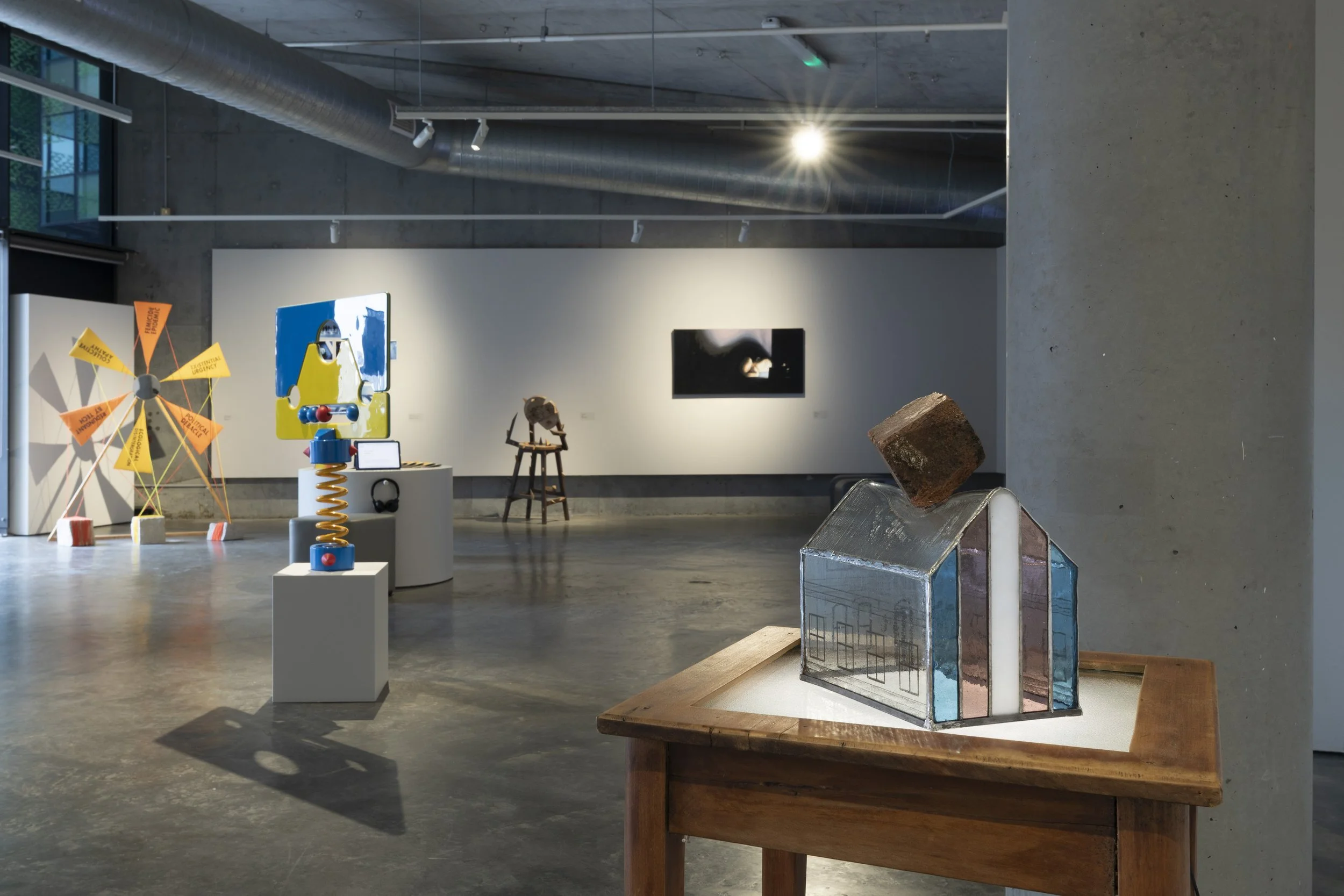Left to right. Children's toy style pillory. Sculpture of a brick on top of a small stained-glass house with trans flag, warping the roof. Sitting on top of a table with light from below.
