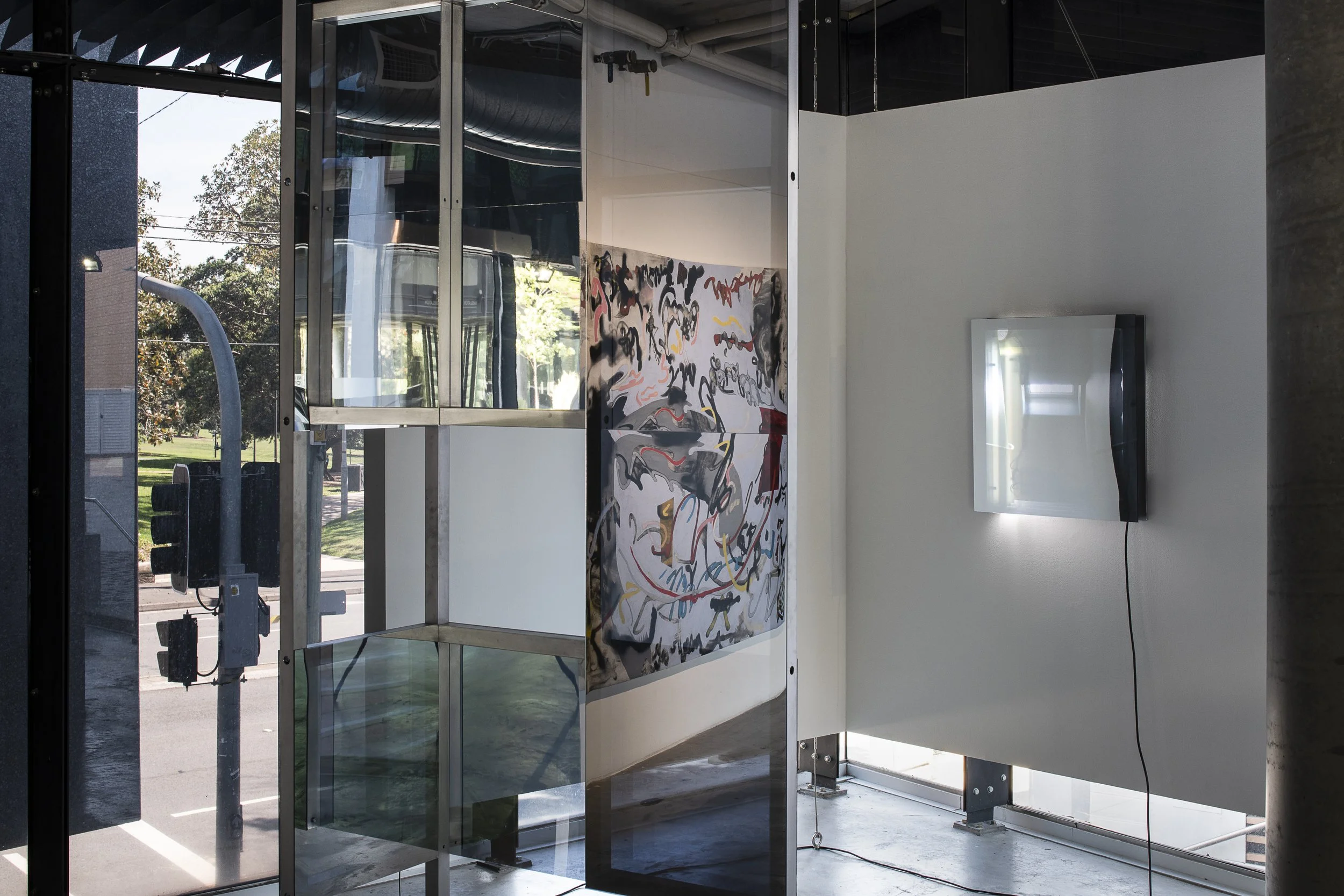Installation view. Left, a large steel frame and mirror structure, reflecting a painting on the left. Right, an acrylic sheet square sculpture on the wall, lit with a fluorescent light from inside.