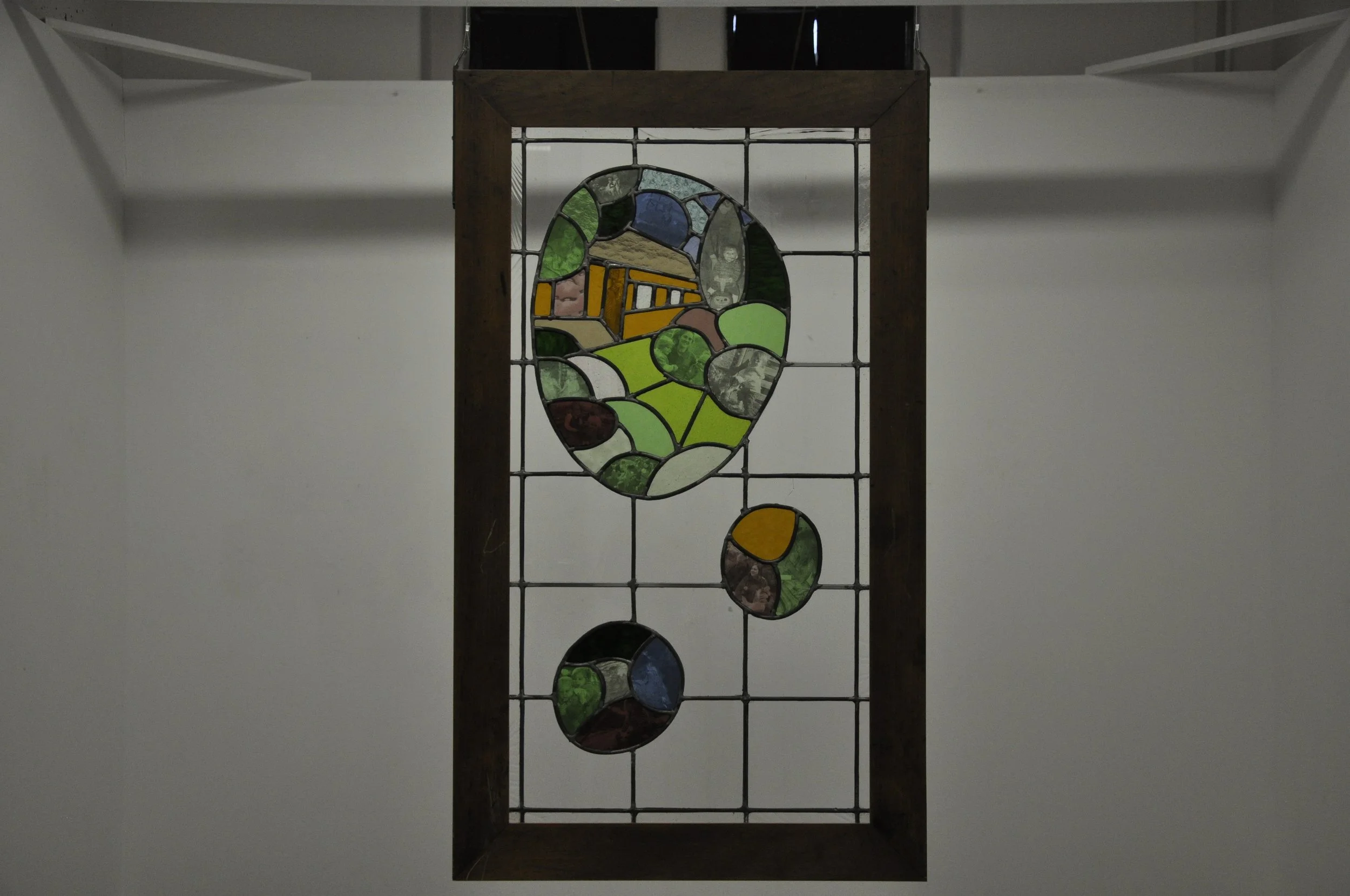 Iris Cuddy, The View Inside, 2026. Wood framed stained glass piece in colours green, orange, grey and blue. The multicoloured glass depicts a house, with transferred images of childhood in on each panel.