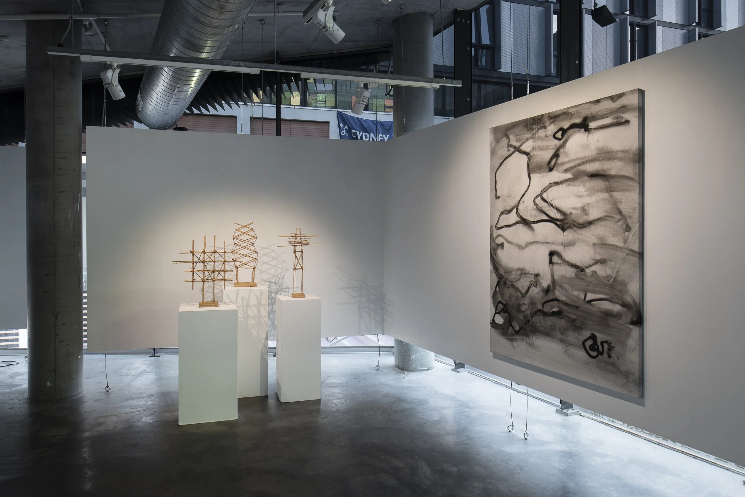 Left, 3 sculptures with structures made of bamboo sticks on 3 plinths. Right, Large abstract painting of smudged black lines on a white background.